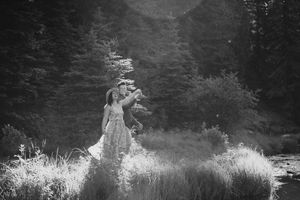 Black and white photo of a couple embracing in a lush meadow surrounded by tall trees and wild grass.