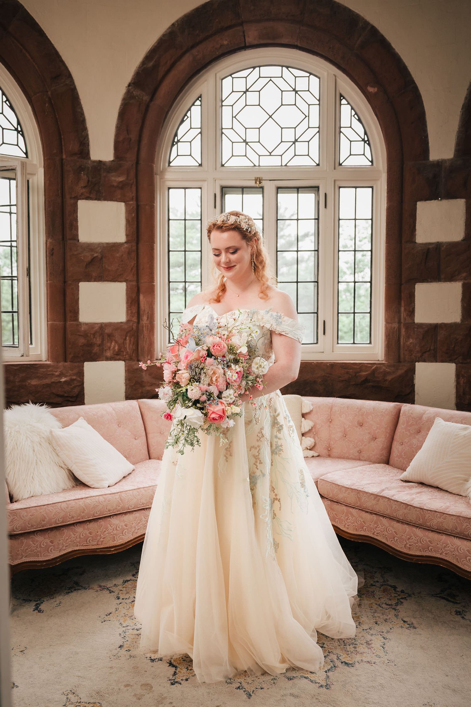Bride looking down at her bouquet in a sunroom with arched stone windows and a pink velvet sofa at Searles Castle.