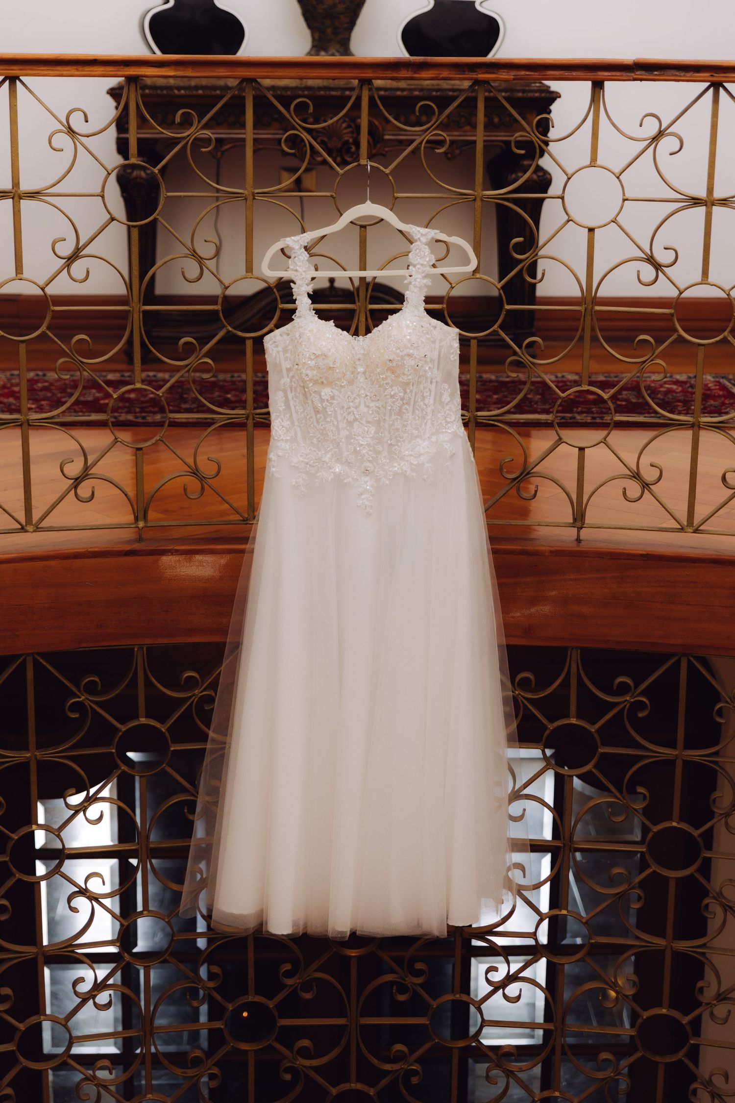 Elegant lace and tulle wedding dress hanging on a hanger against a decorative golden iron staircase railing.