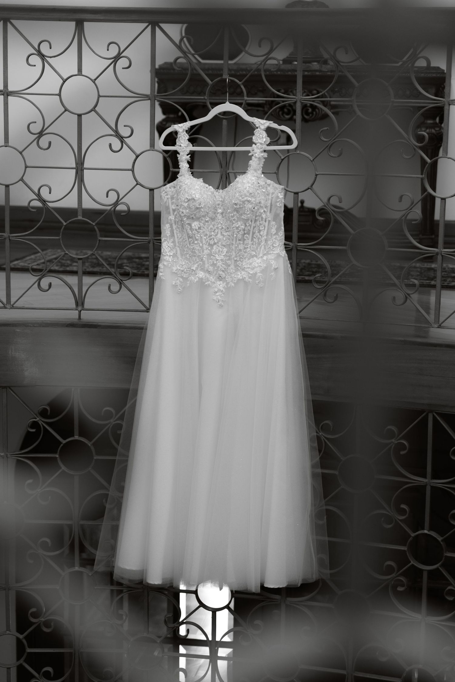 Black and white photo of a lace and tulle wedding gown hanging on an ornate iron railing.