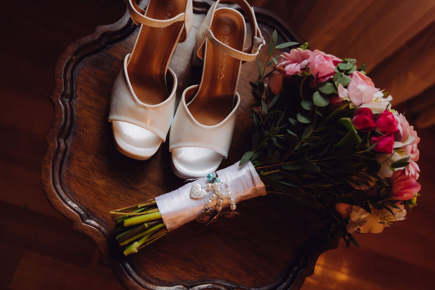 White platform heels and a pink and rose bridal bouquet with pearl charm displayed on a wooden table.