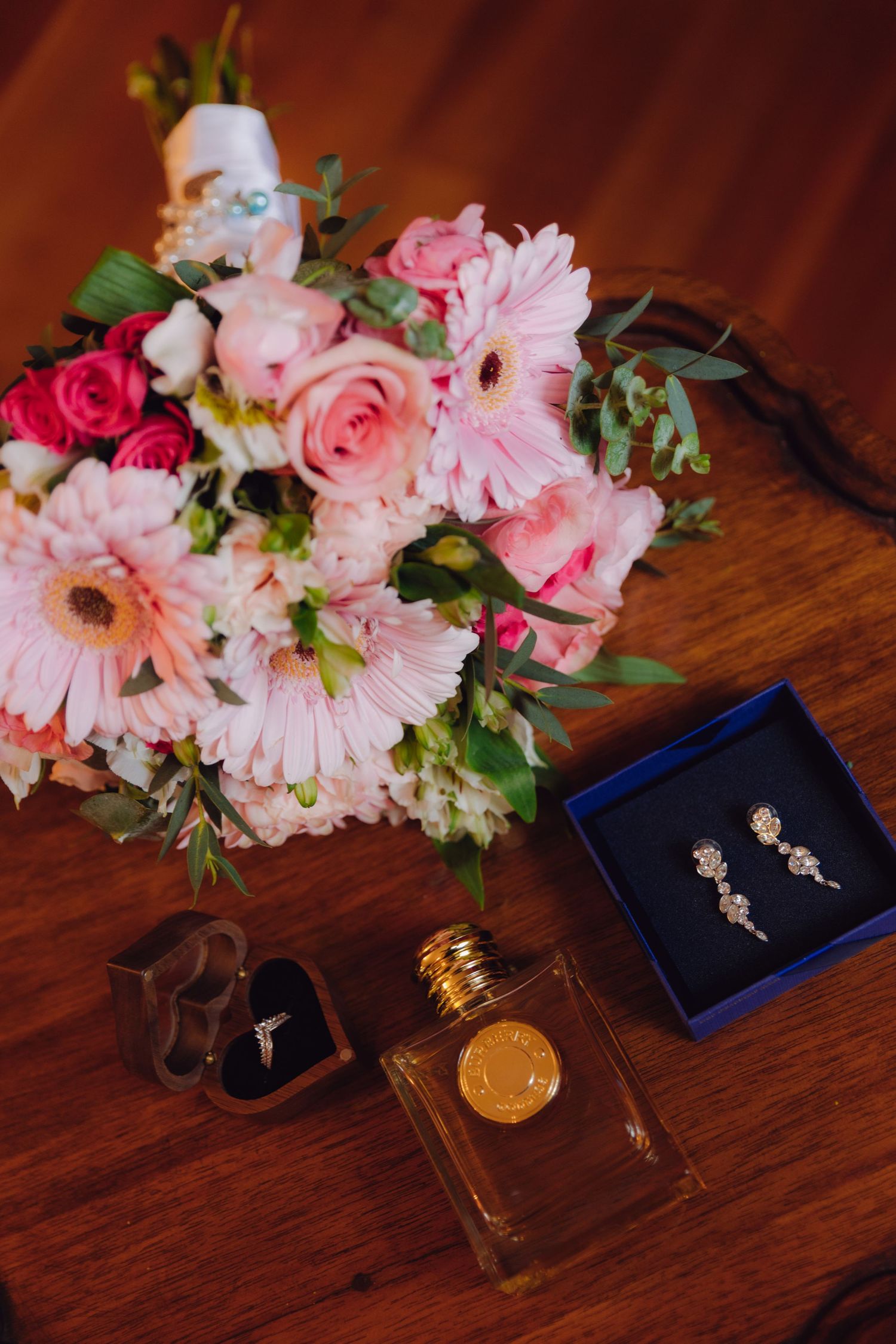 Pink bridal bouquet with gerberas and roses beside diamond earrings, perfume, and a ring box on wood.