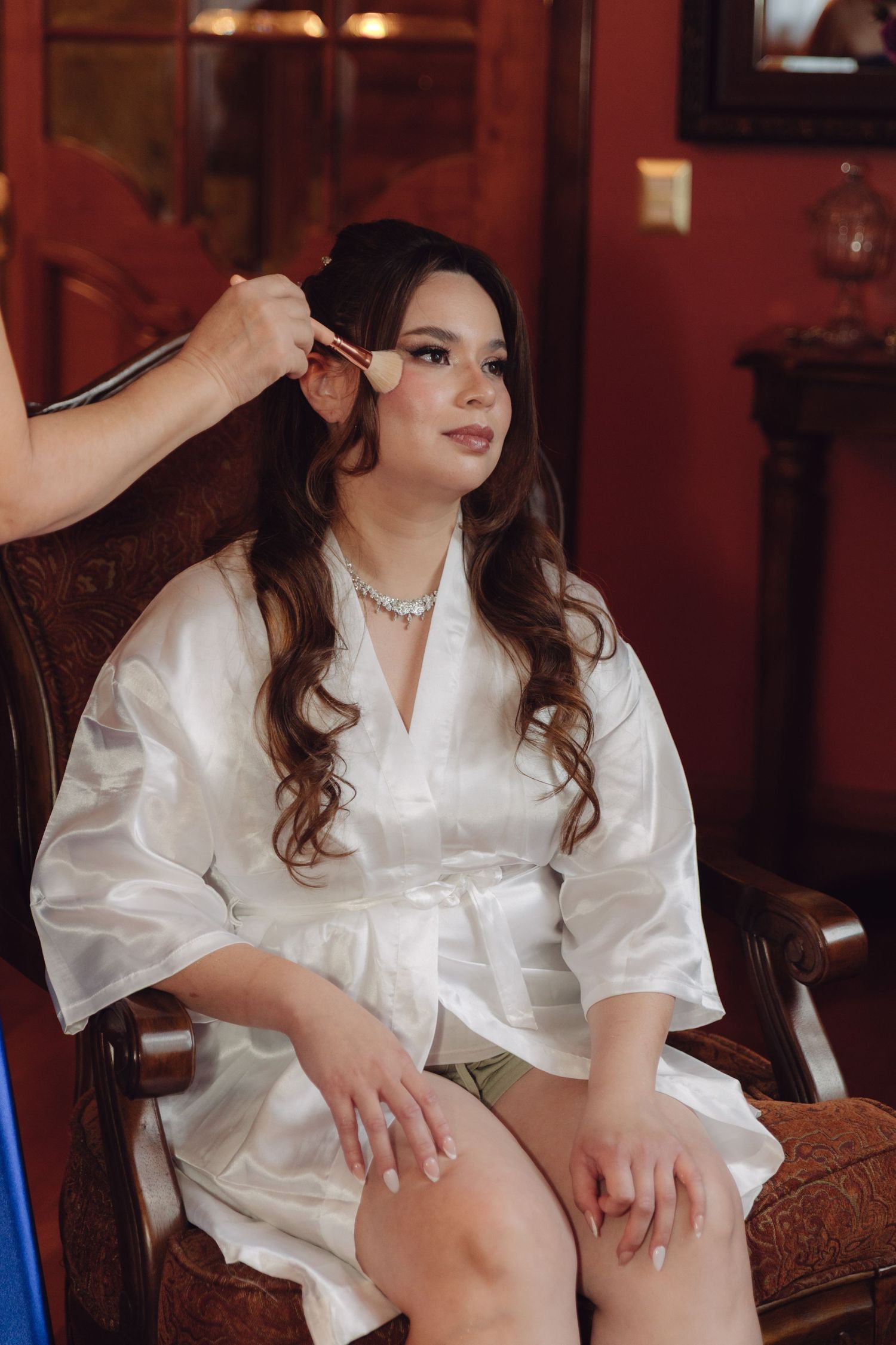 Bride in white satin robe getting makeup applied with a brush while seated in an ornate wooden chair.