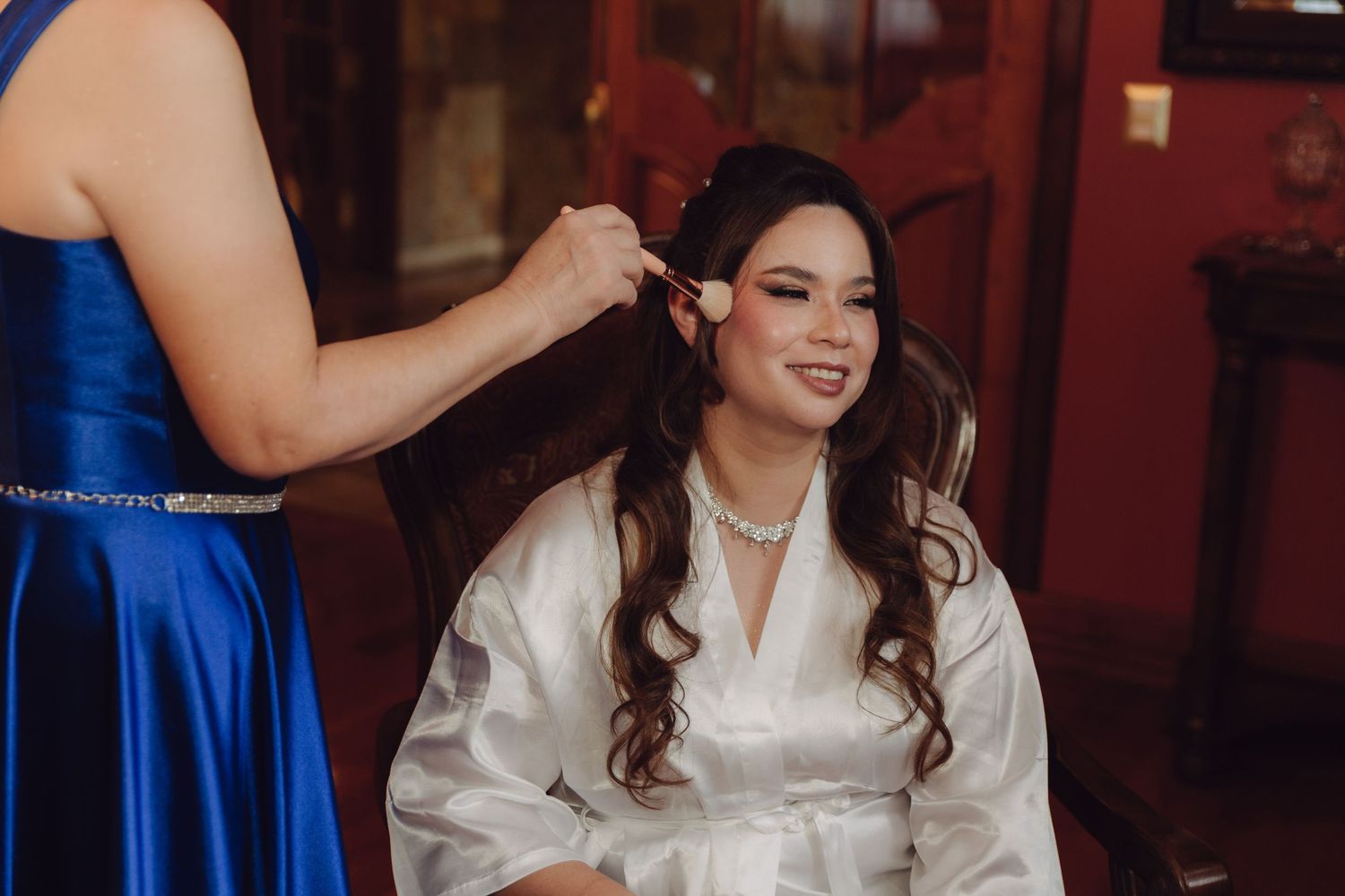 Smiling bride in white robe receiving makeup application from a woman in a blue satin dress.