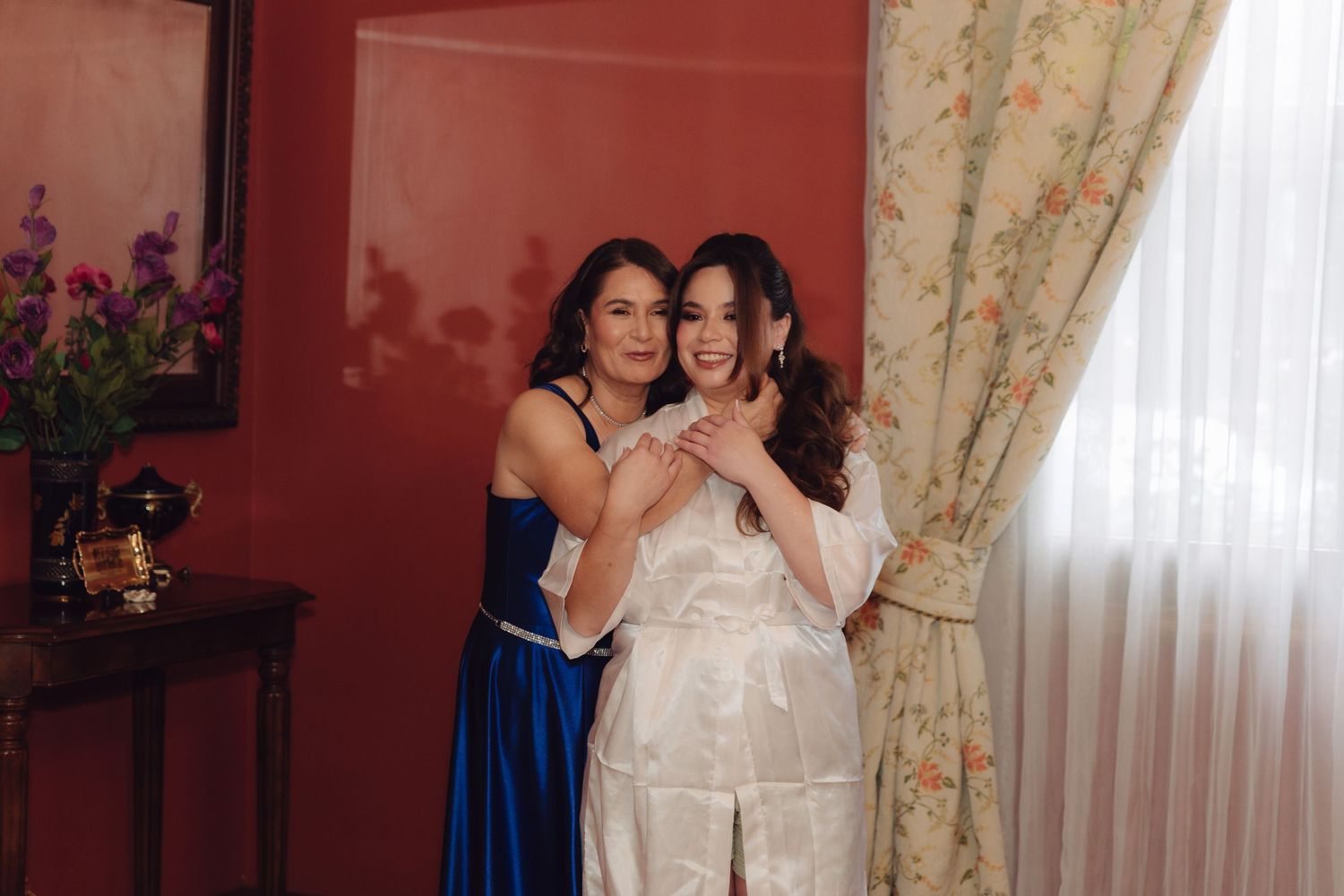 Bride in white satin robe embracing a woman in a royal blue dress in an elegantly decorated room.