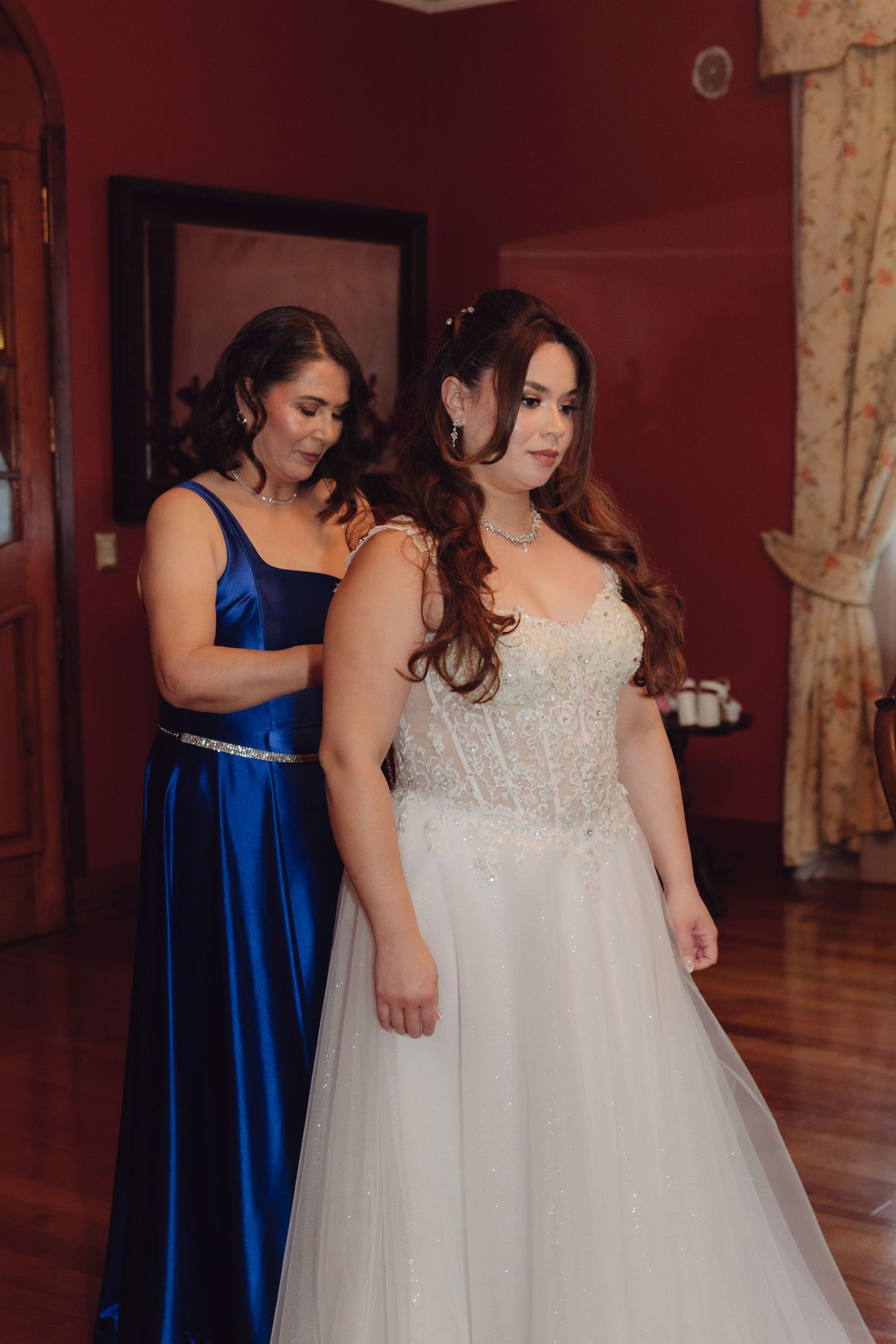 Woman in royal blue gown helping a bride in a beaded white wedding dress in a red-walled room.