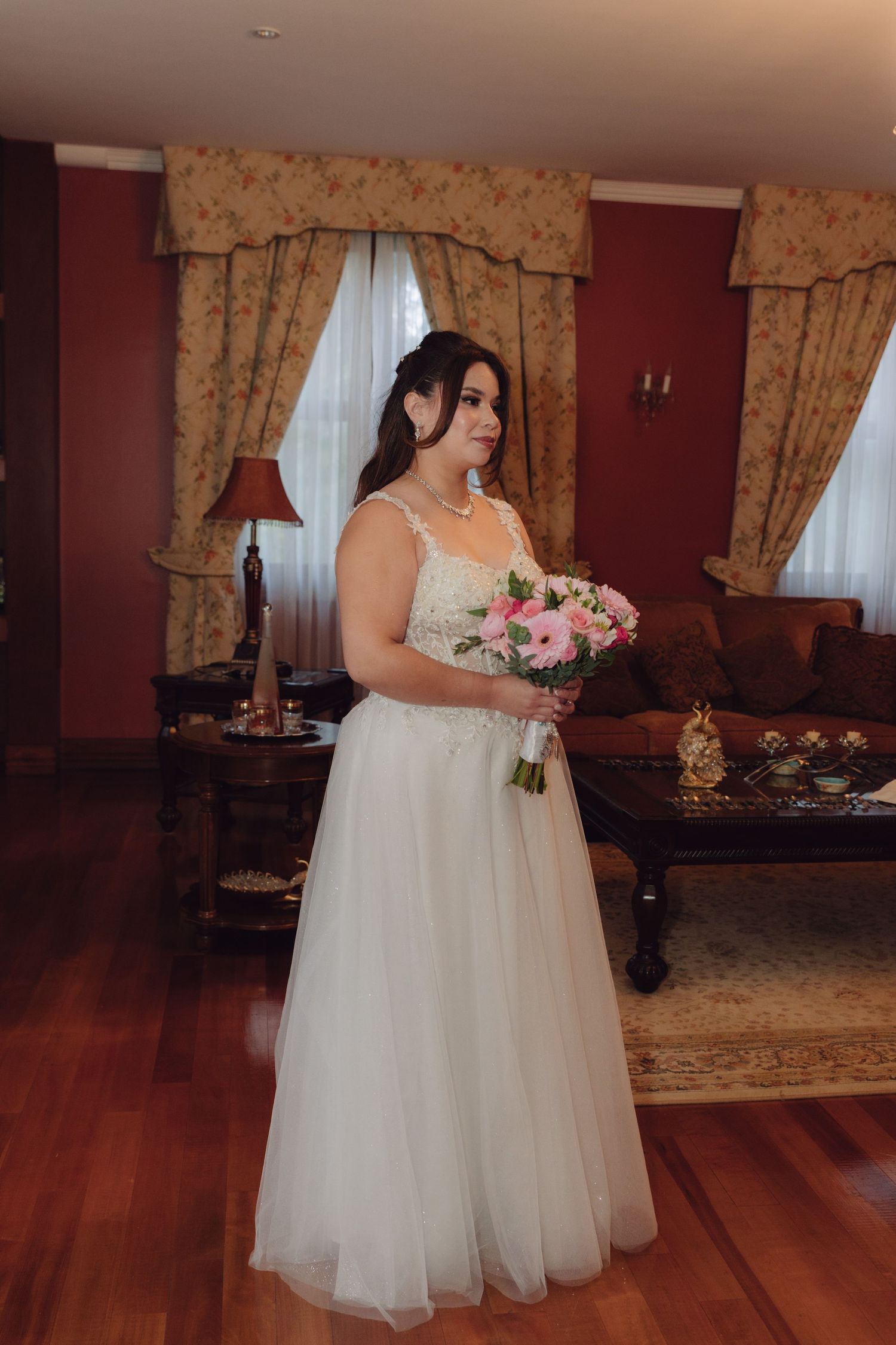 Bride in a white lace and tulle wedding gown holding a pink bouquet standing in an elegant room.