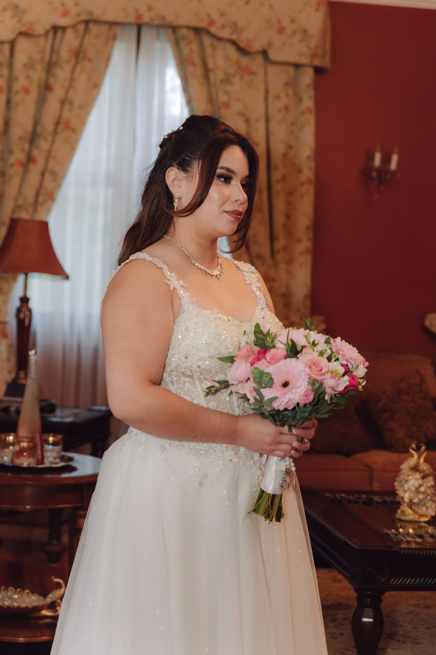 Bride wearing a beaded white wedding gown holds a pink and white bouquet in a warmly lit room.