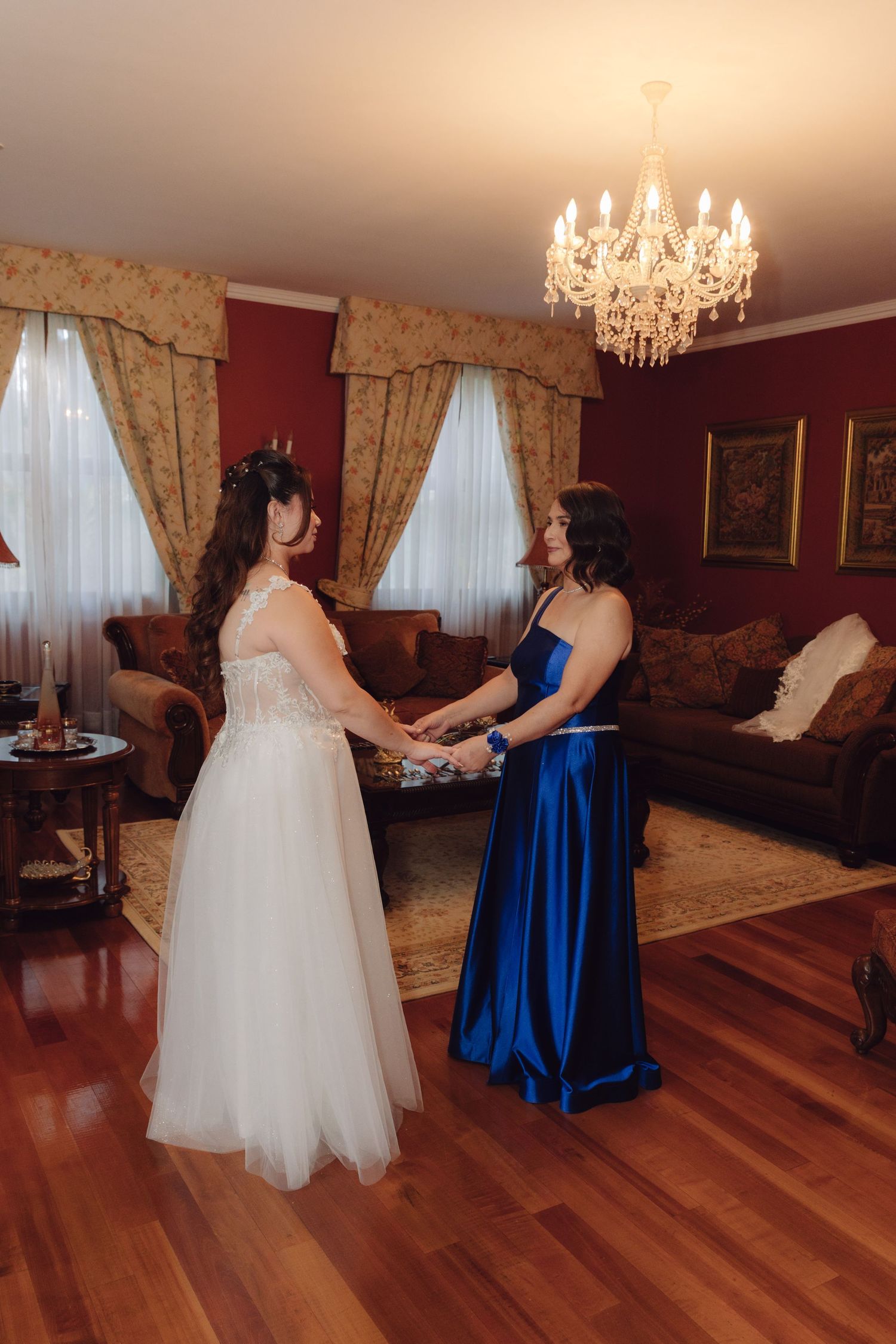 Bride in white gown and woman in royal blue dress holding hands under a crystal chandelier.