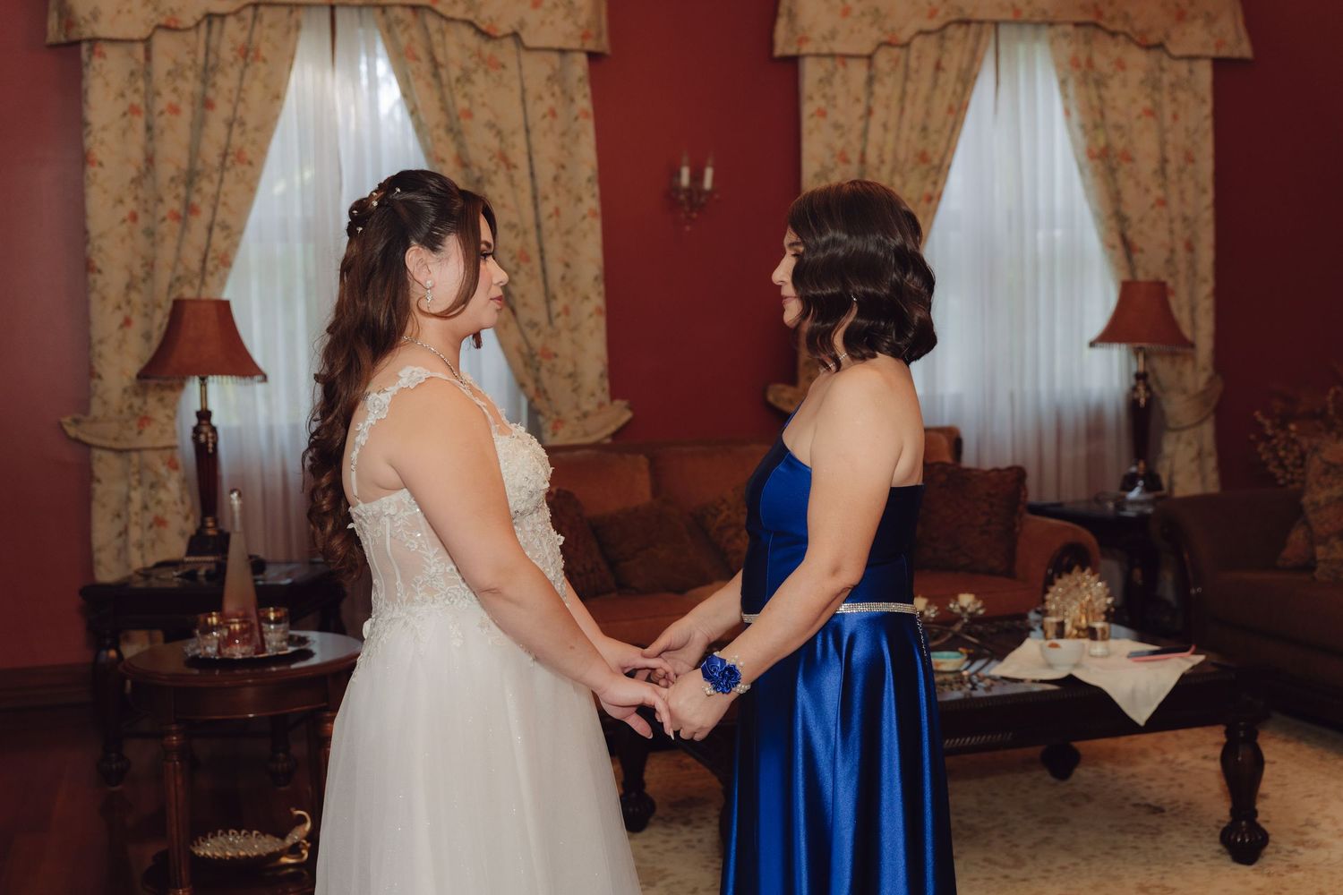 Bride in white wedding dress and woman in royal blue gown holding hands facing each other indoors.