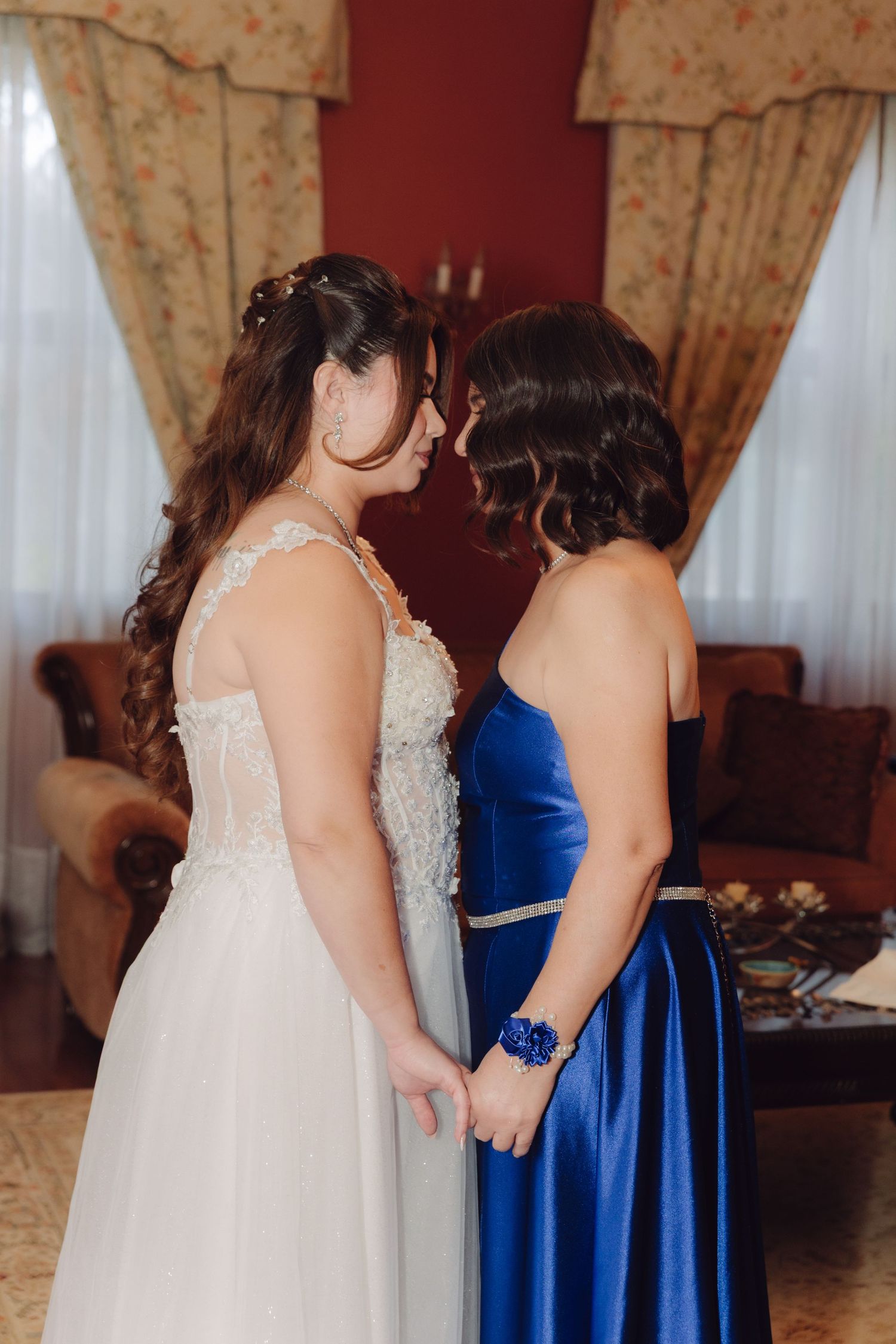 Bride in white lace gown and companion in blue satin dress holding hands facing each other closely.