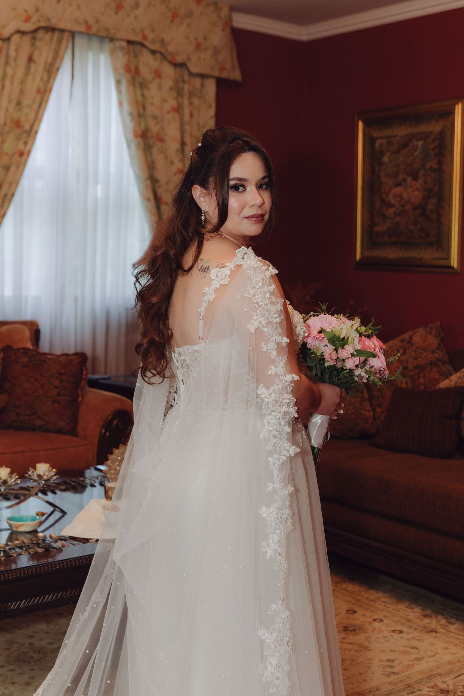 Bride in elegant white lace wedding gown with sheer cape holding pink bouquet in ornate red room.