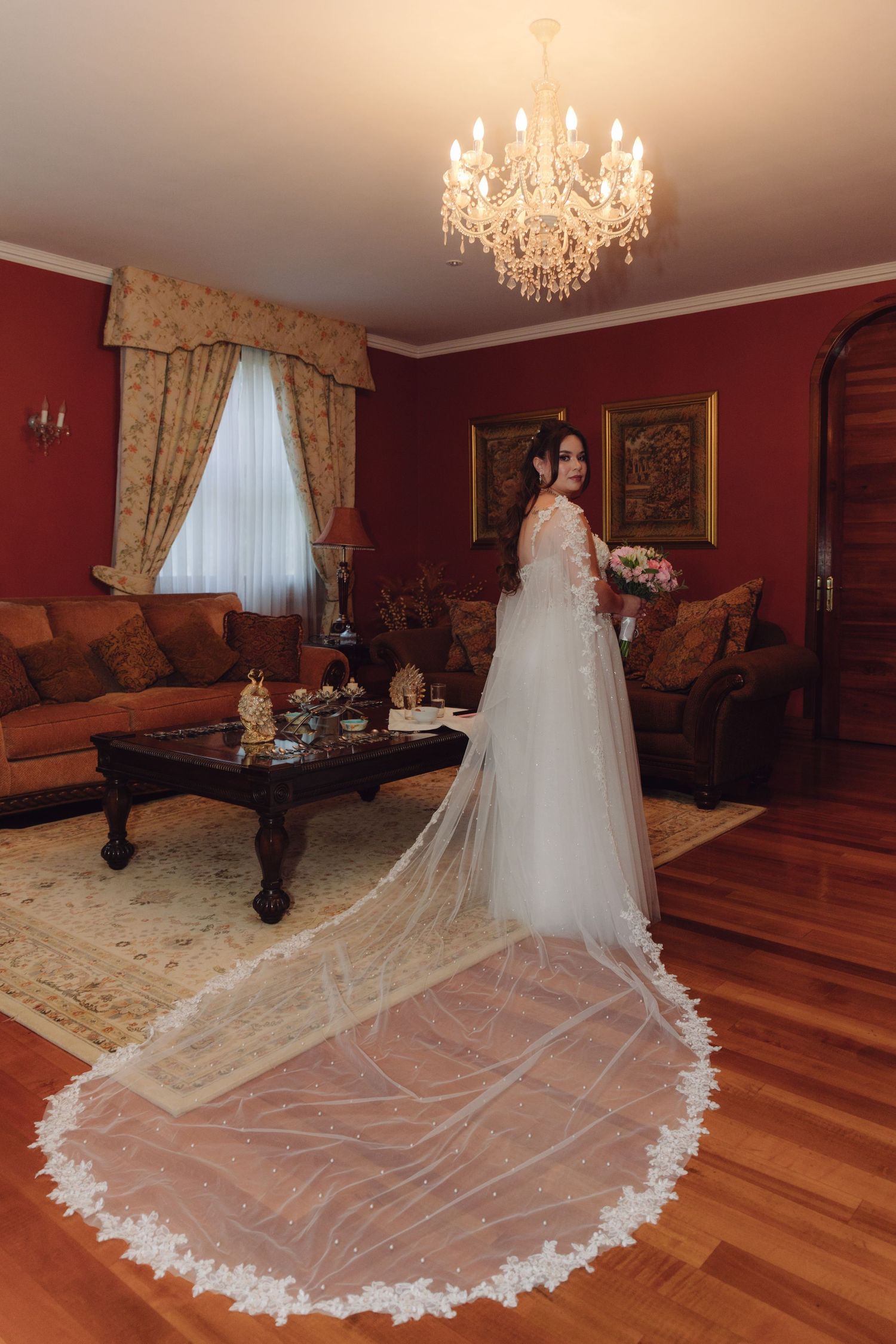 Bride in stunning white gown with long lace-trimmed cathedral veil standing in elegant red living room.