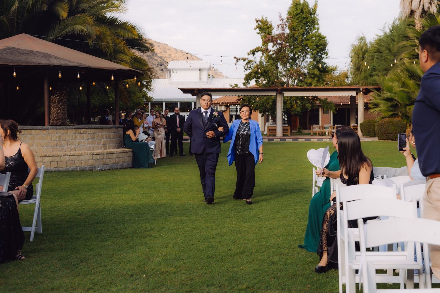 Groom escorted by woman in blue jacket walking down outdoor wedding ceremony aisle on green lawn.