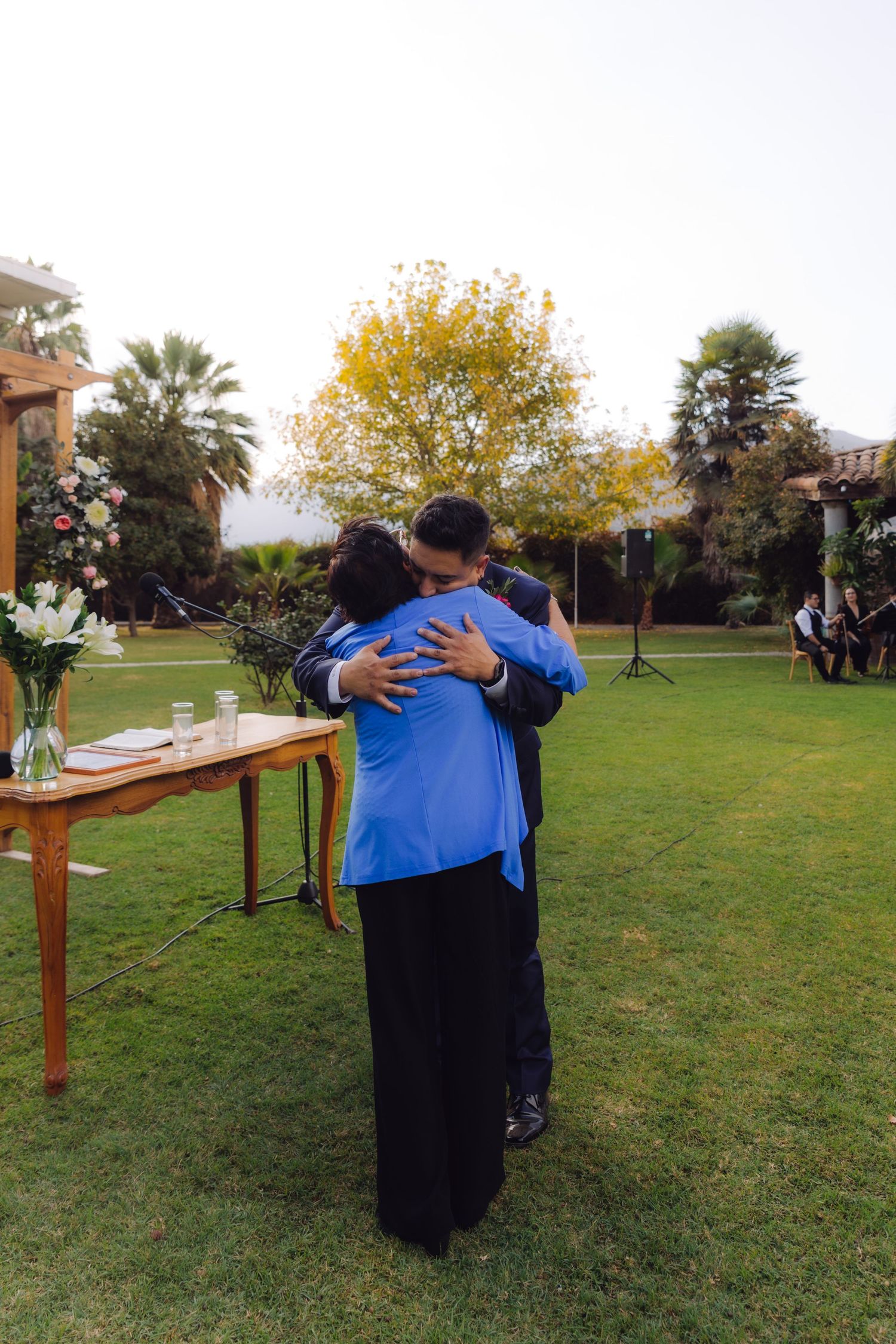 Groom in navy suit sharing emotional hug with woman in blue jacket at outdoor wedding ceremony.