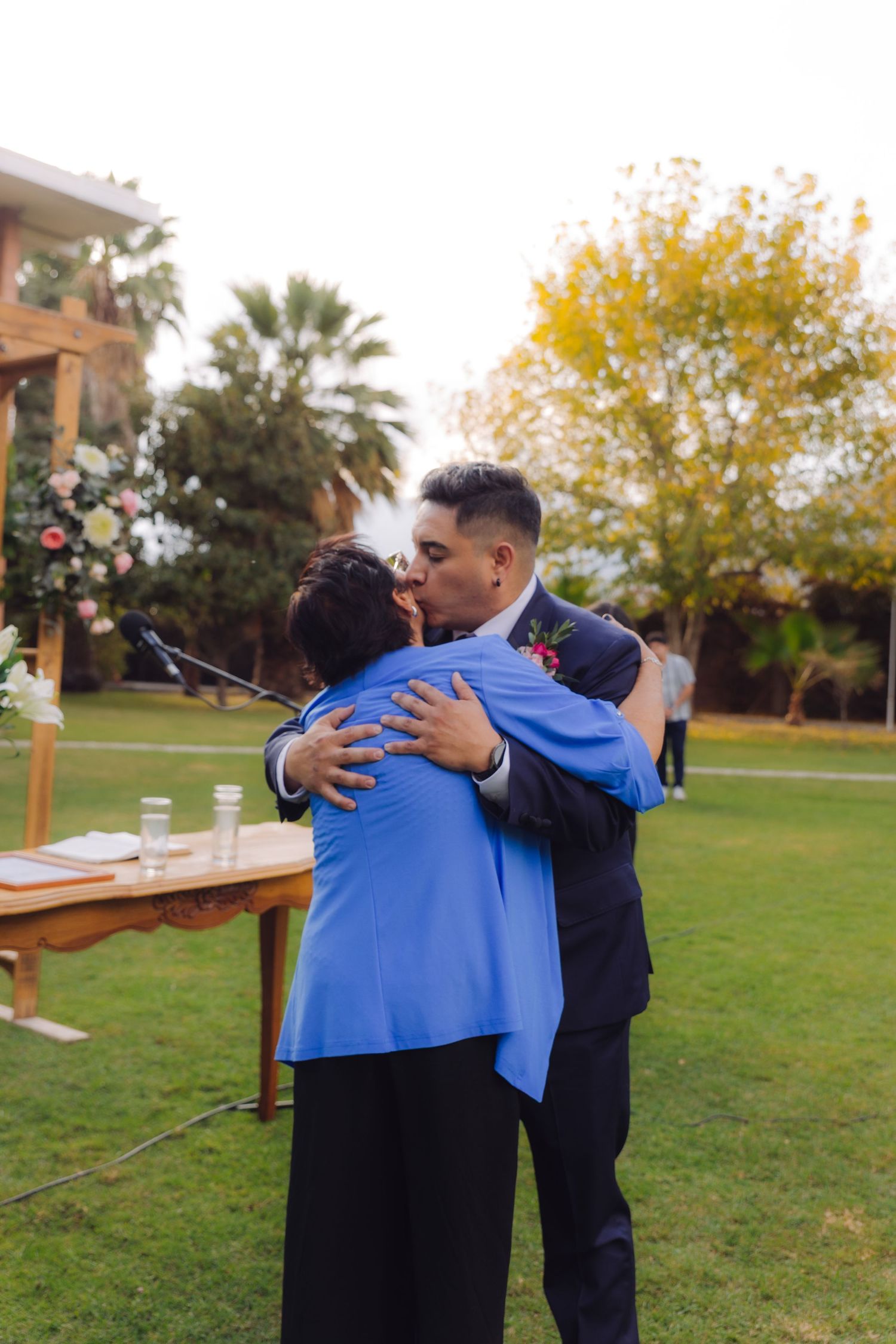 Groom in navy suit kissing woman in blue jacket on cheek at outdoor garden wedding ceremony.