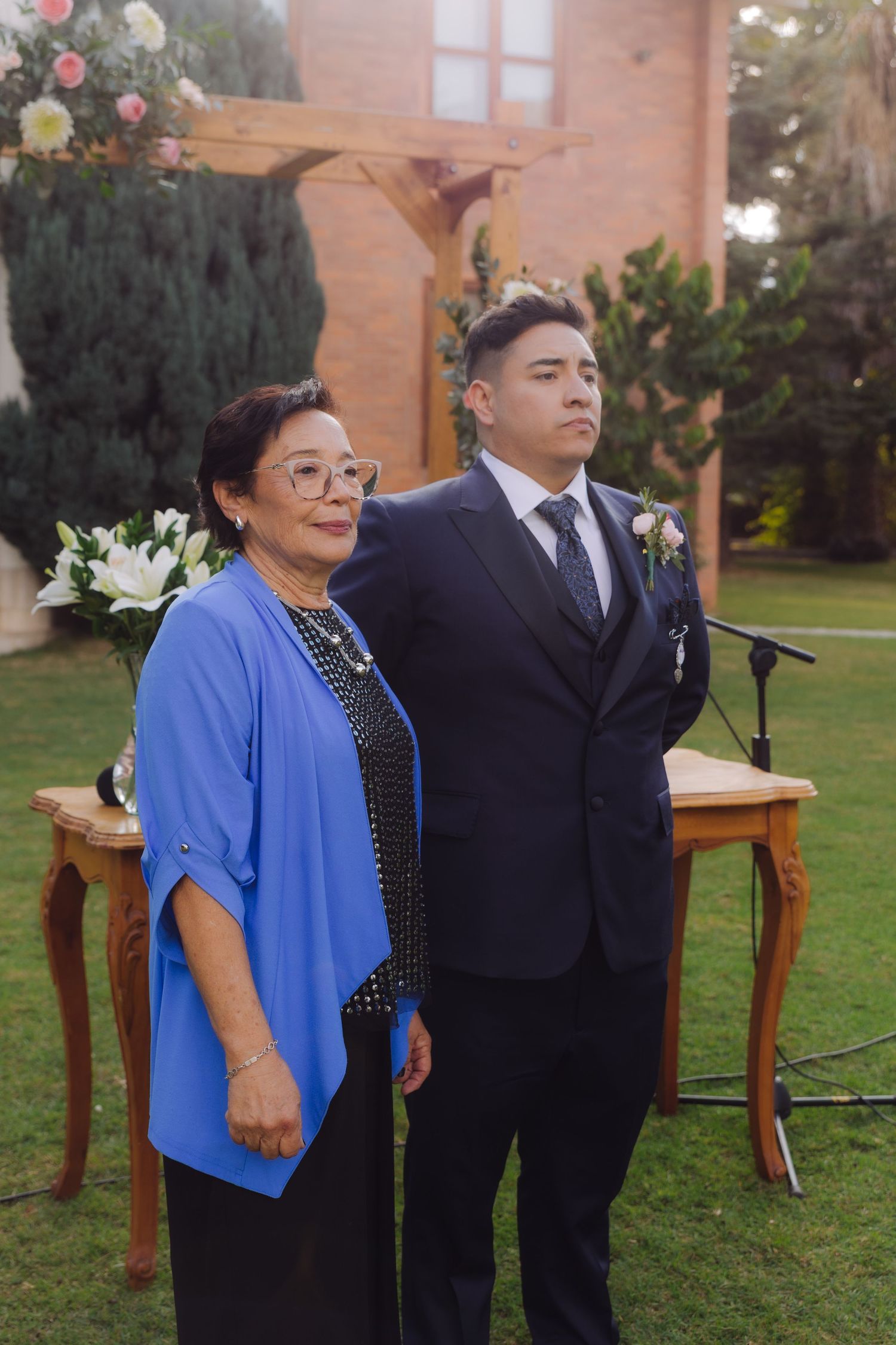 Groom in navy suit with boutonniere standing beside older woman in blue jacket at outdoor altar.