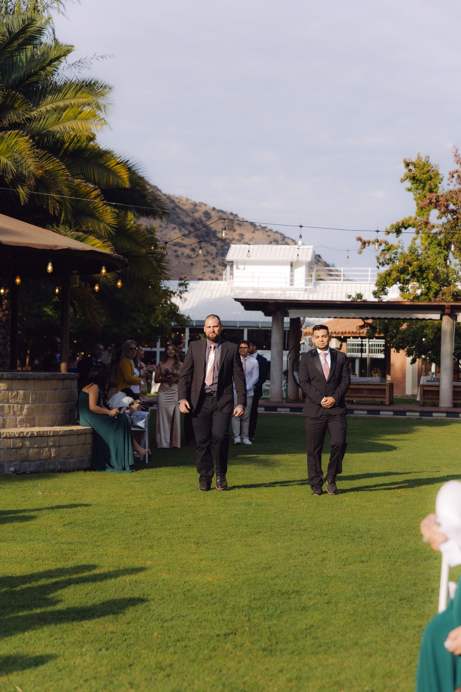 Two groomsmen in dark suits with pink ties walking across green lawn at outdoor wedding venue.