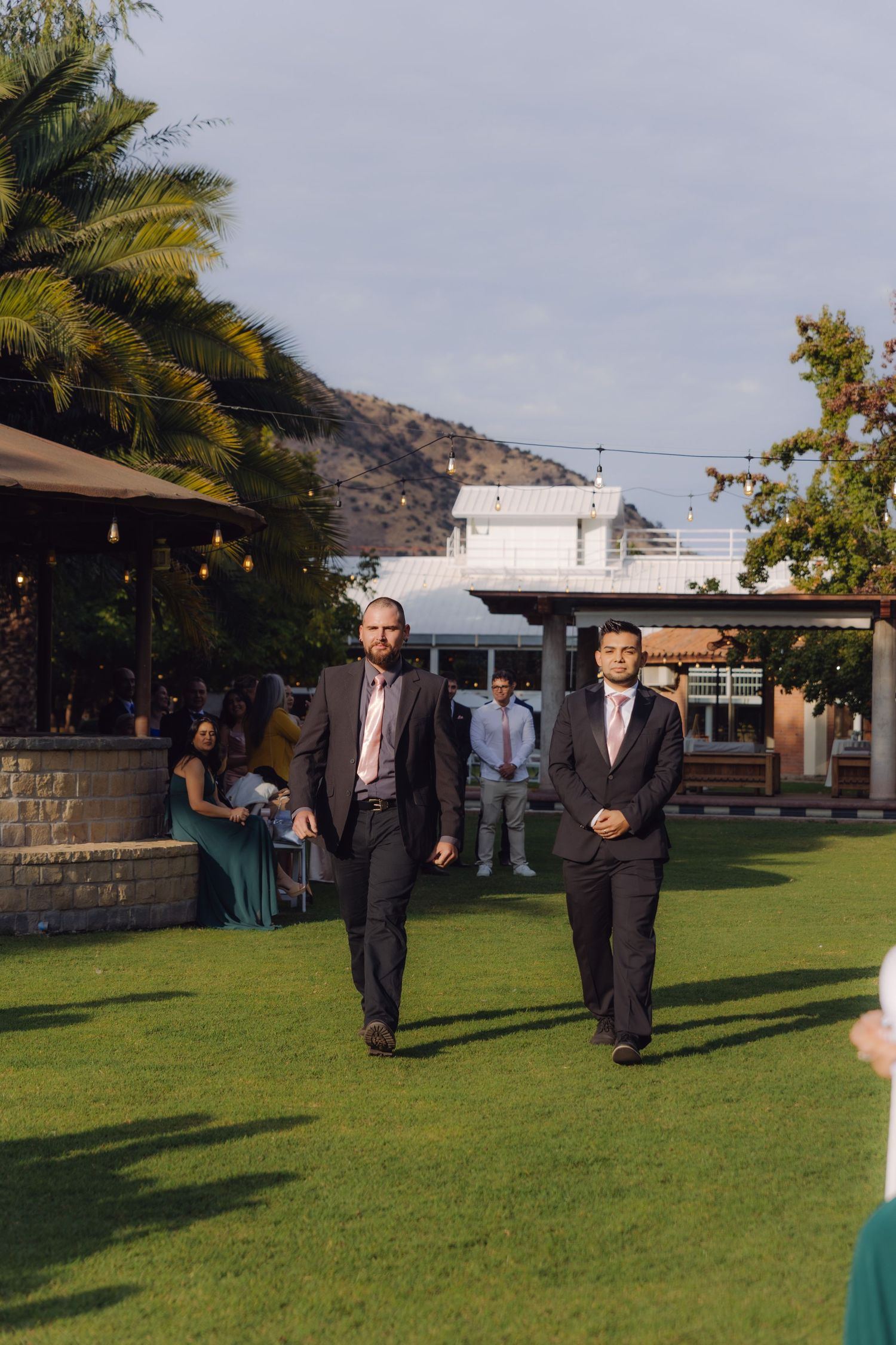 Two groomsmen in dark suits with pink ties walking across manicured lawn at outdoor wedding ceremony.