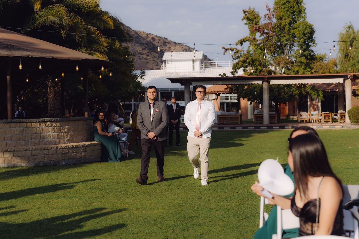 Groomsman in gray suit and groom in white suit walking down outdoor wedding ceremony aisle on lawn.