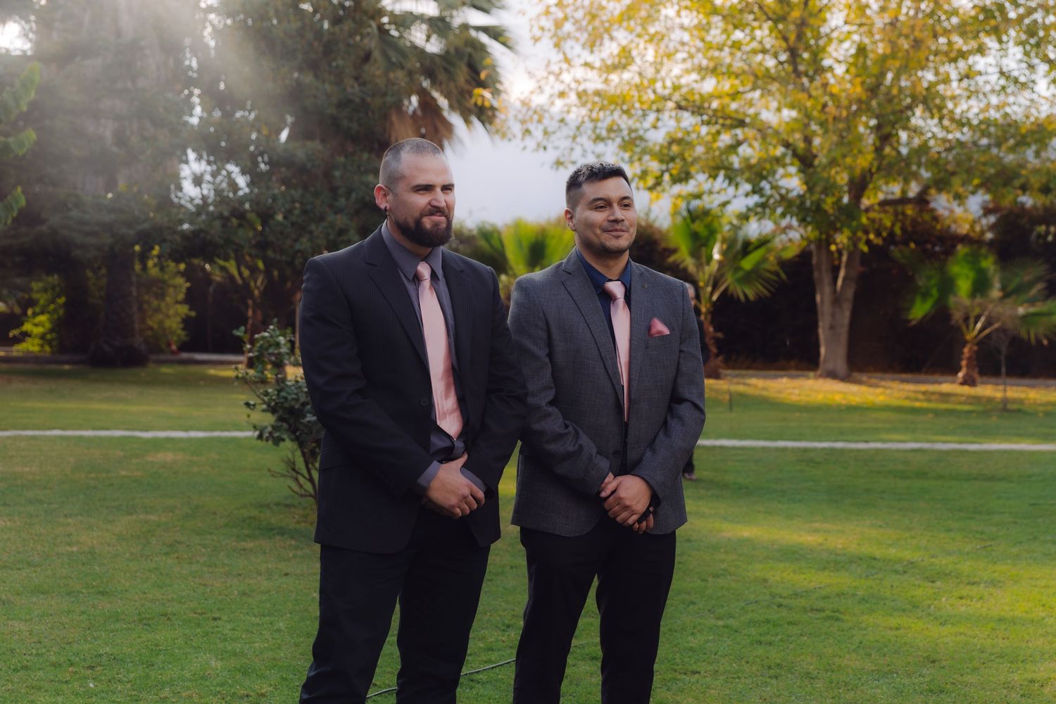 Two groomsmen in dark gray suits with pink ties posing together on green lawn at wedding.