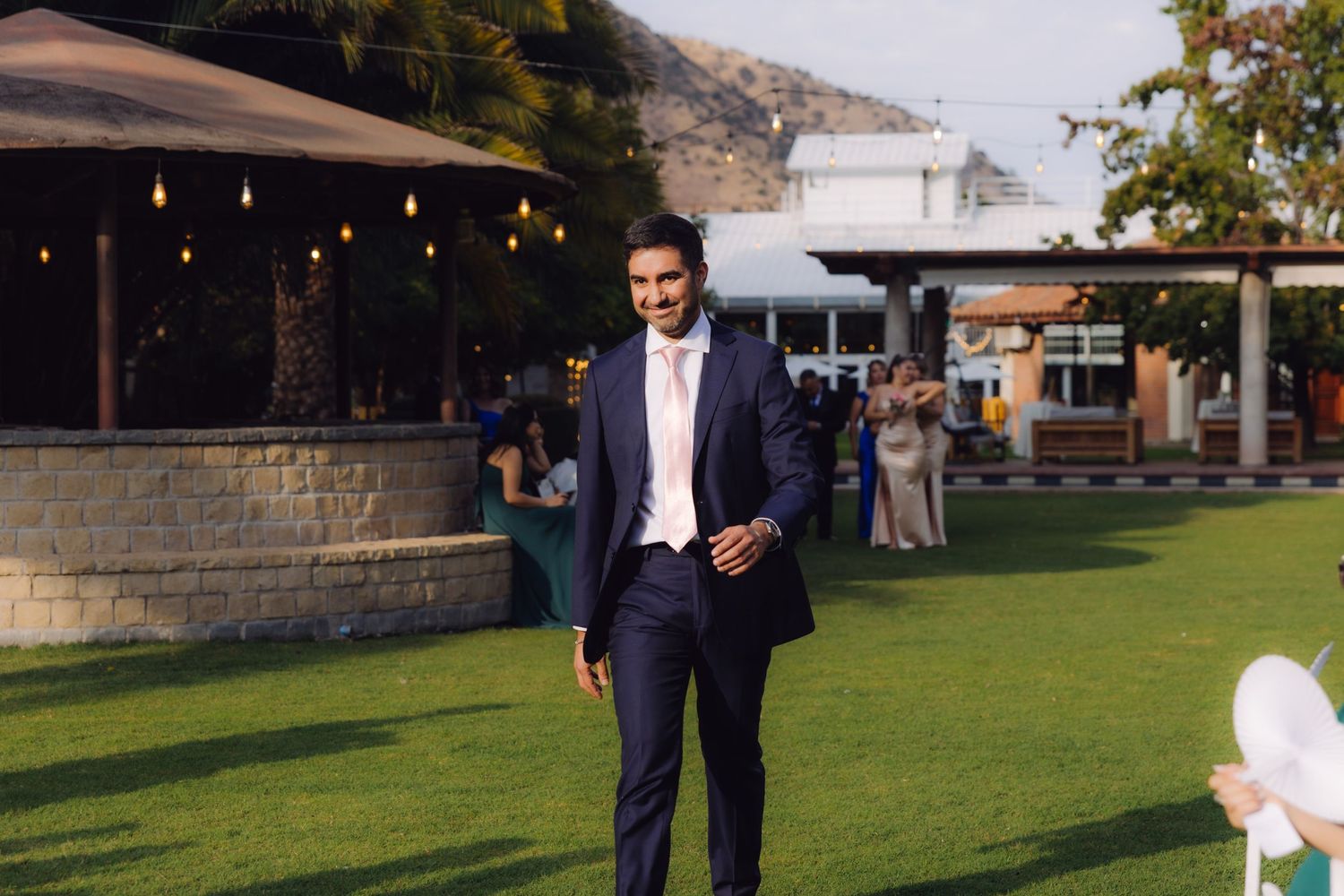 Tall man in navy blue suit walking across green lawn at outdoor wedding venue with mountain backdrop.