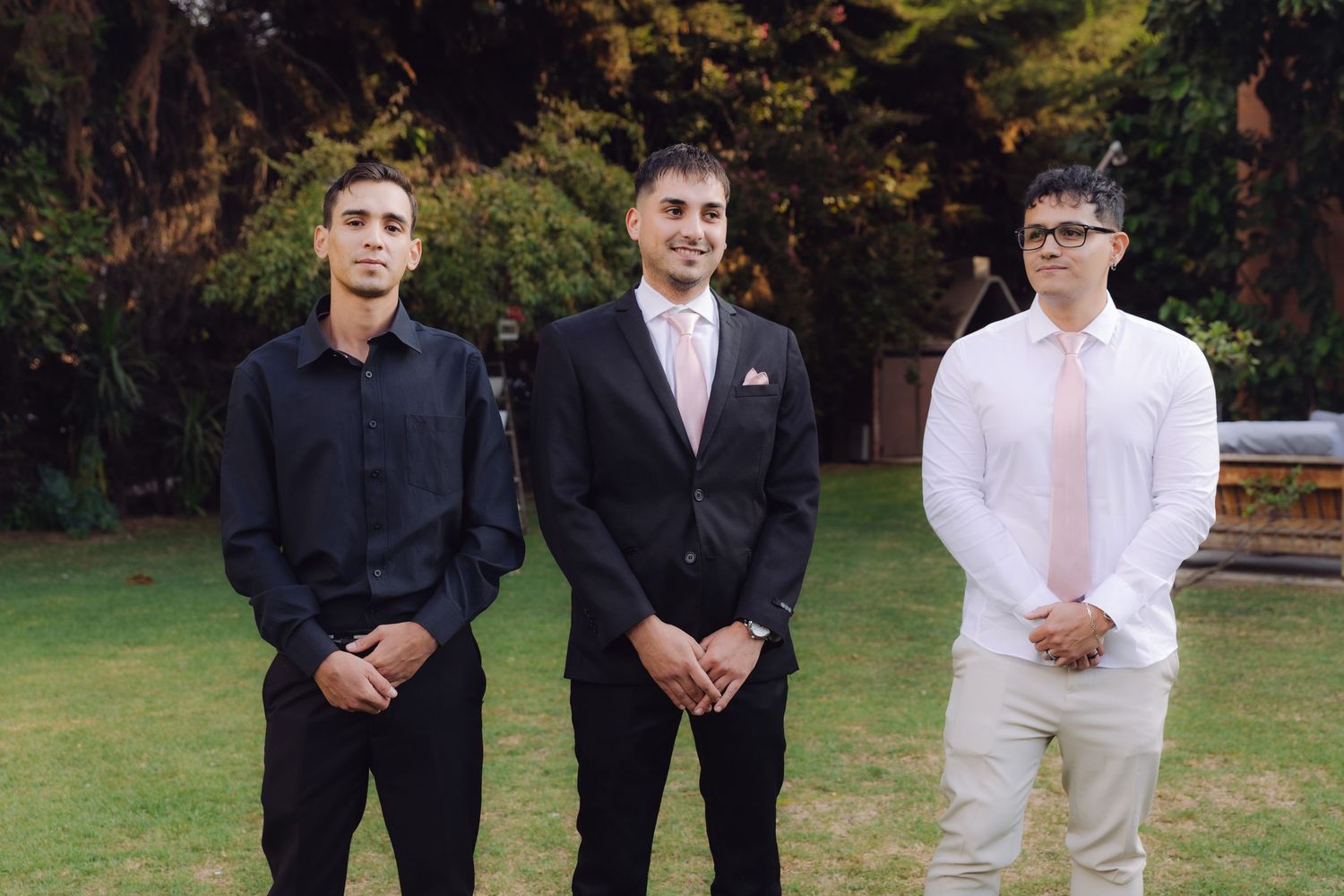 Three young men in formal suits standing together on grass at an outdoor wedding ceremony.