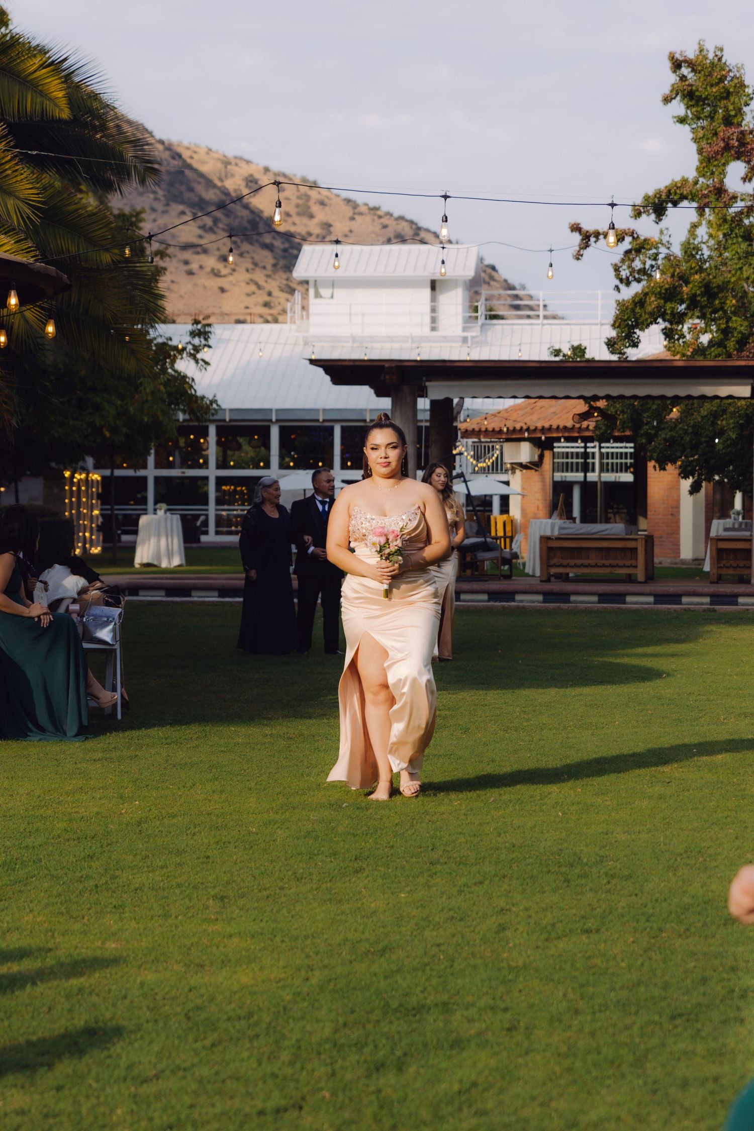 Bridesmaid in champagne satin gown walking down outdoor aisle carrying pink bouquet at wedding.