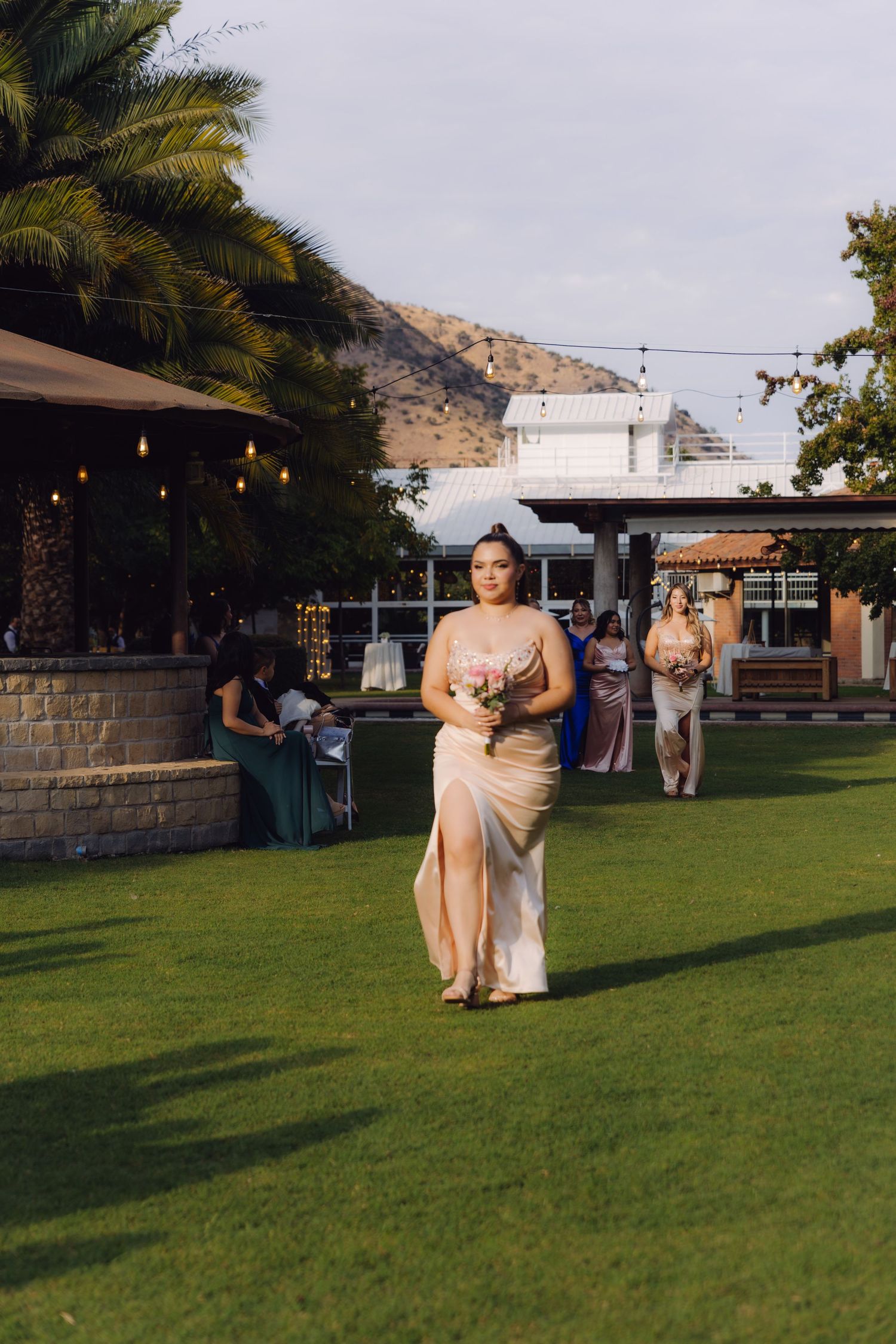Bridesmaid in gold satin dress walking across lawn with mountain and venue building in background.