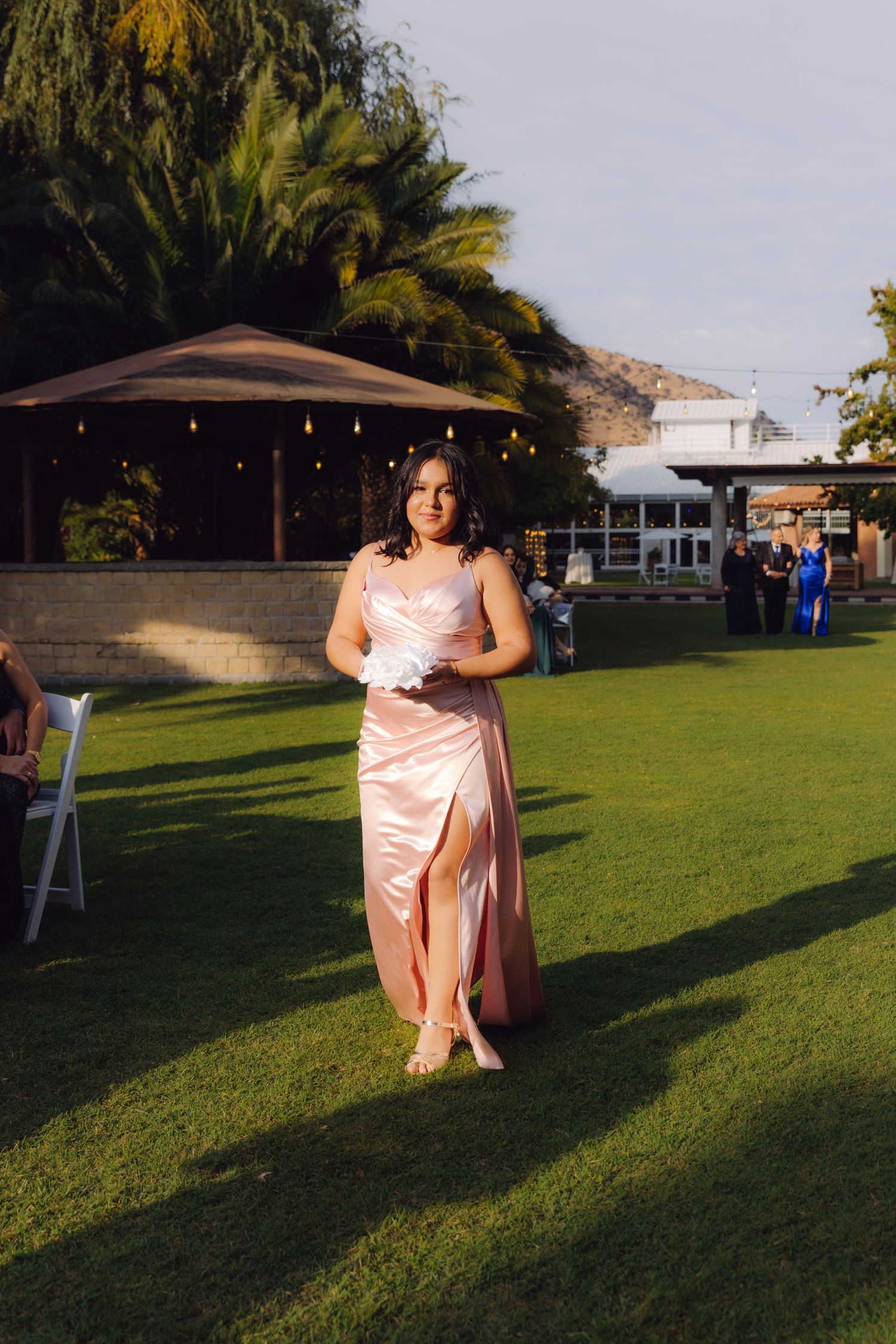Young woman in blush pink satin bridesmaid dress holding bouquet walking on green grass at wedding.