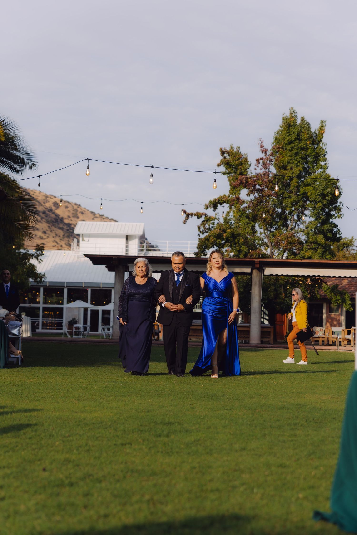 Three elegantly dressed wedding guests walking across lawn in navy and royal blue formal attire.