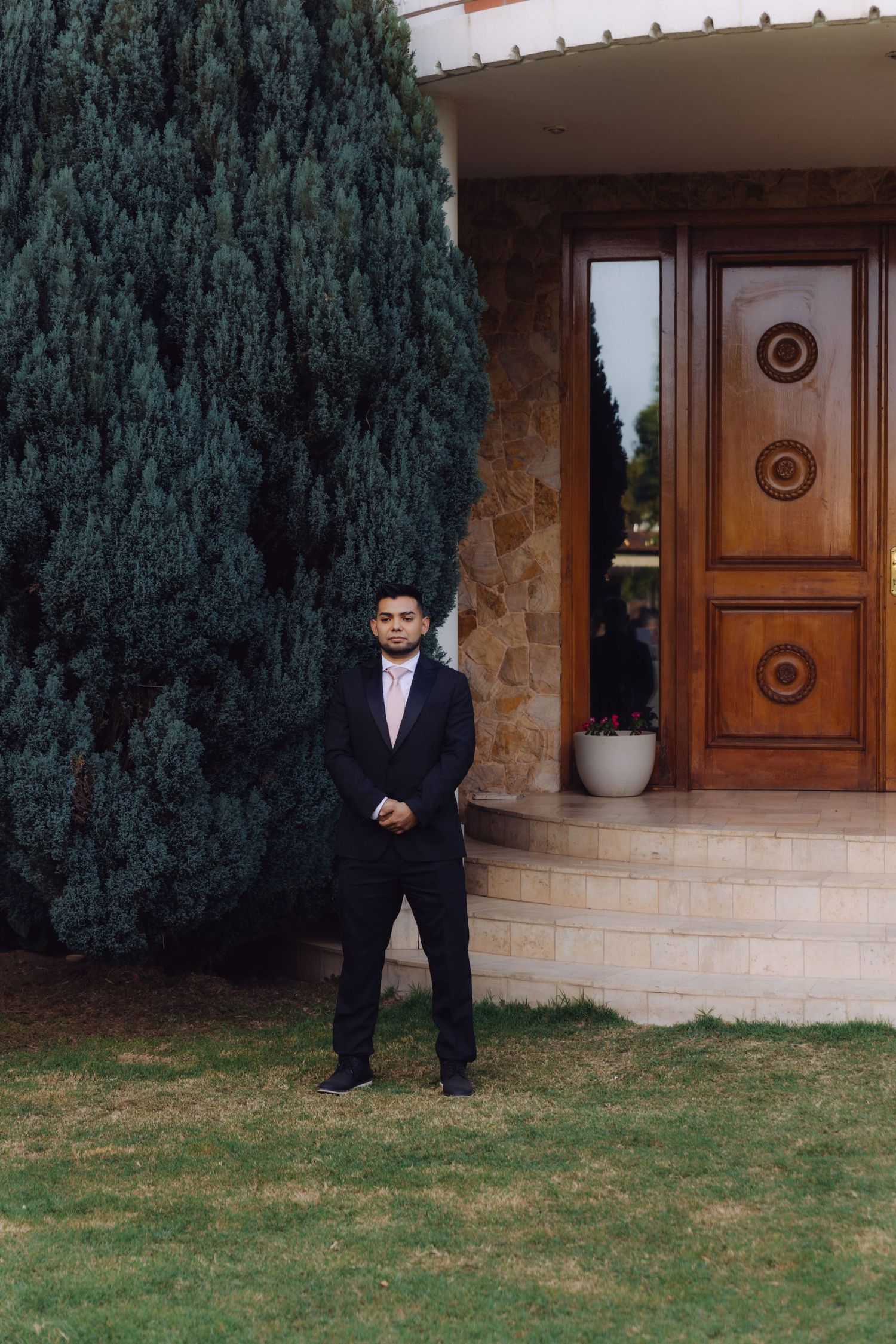 Groom in black suit standing on green lawn beside tall cypress tree near wooden entrance doors.