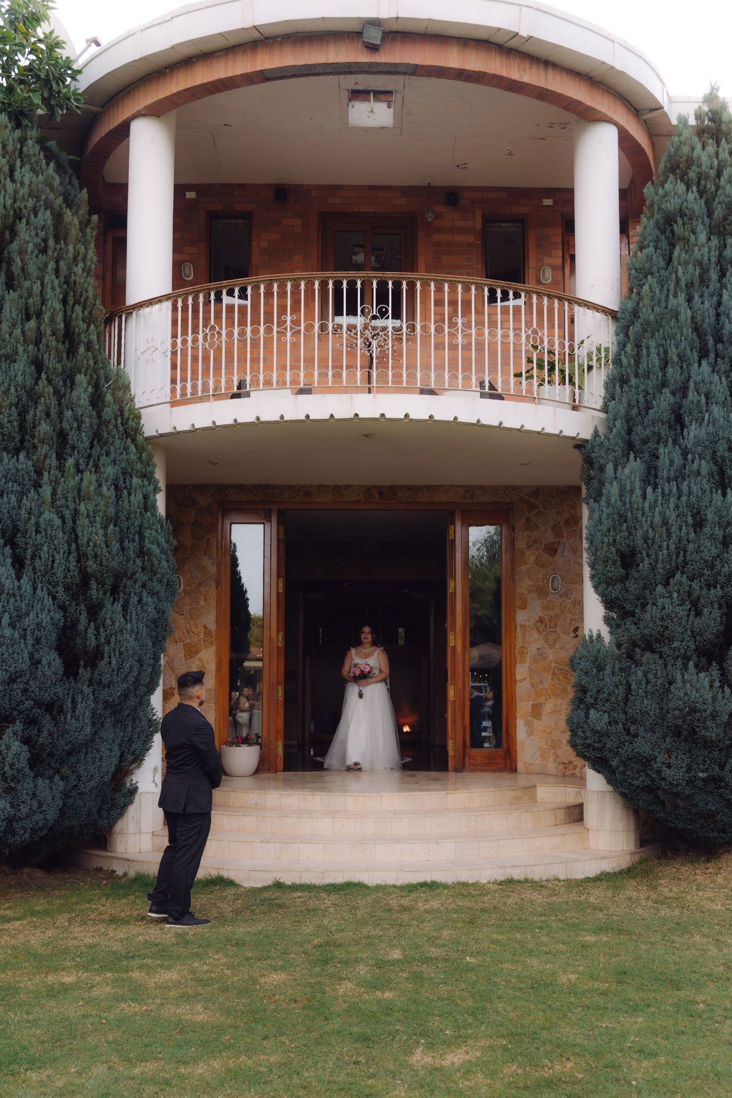 Groom watching bride in white wedding dress descend stone steps of elegant venue entrance.