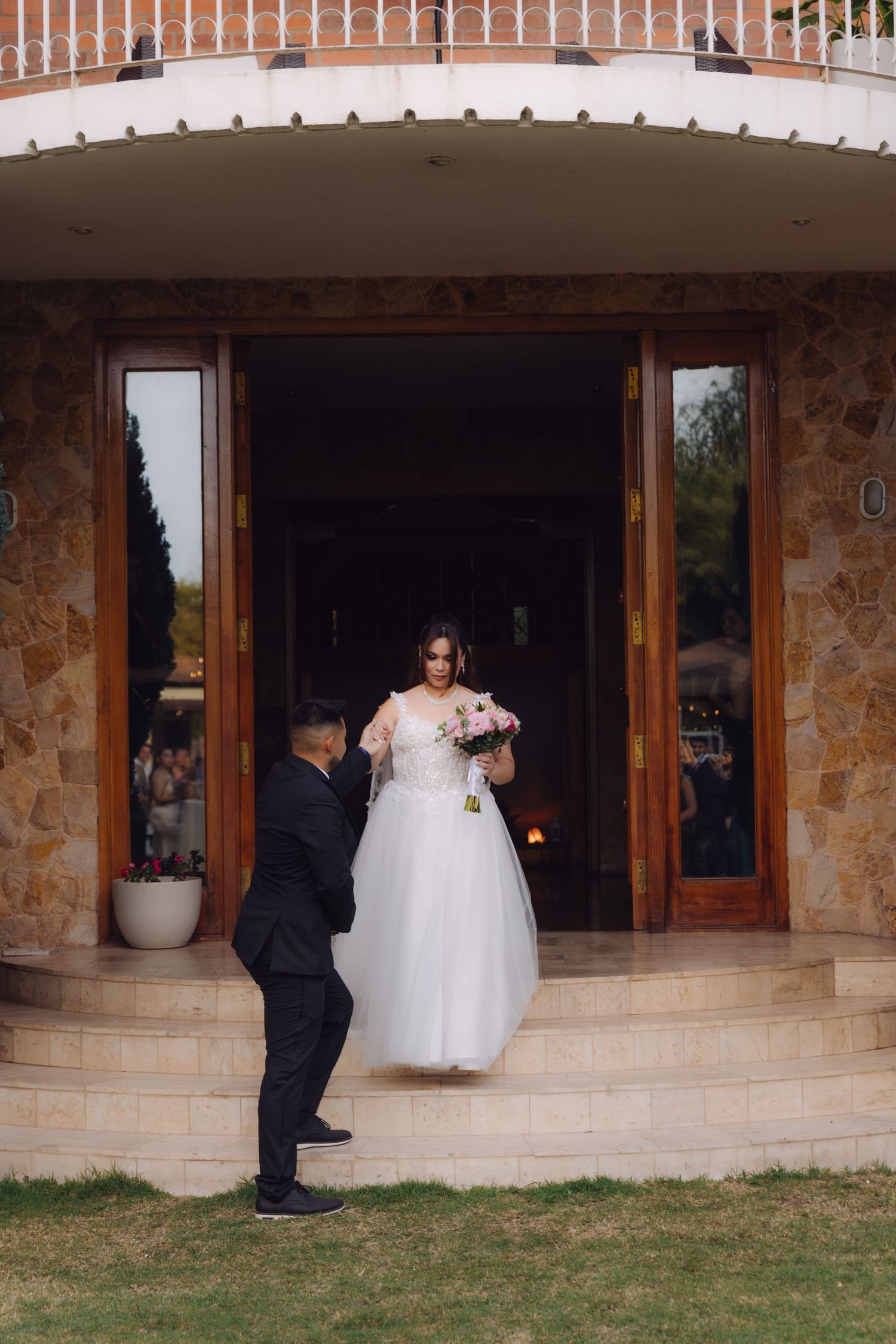 Groom helps bride descend stone steps at wedding venue entrance, bride holds pink bouquet in white gown.