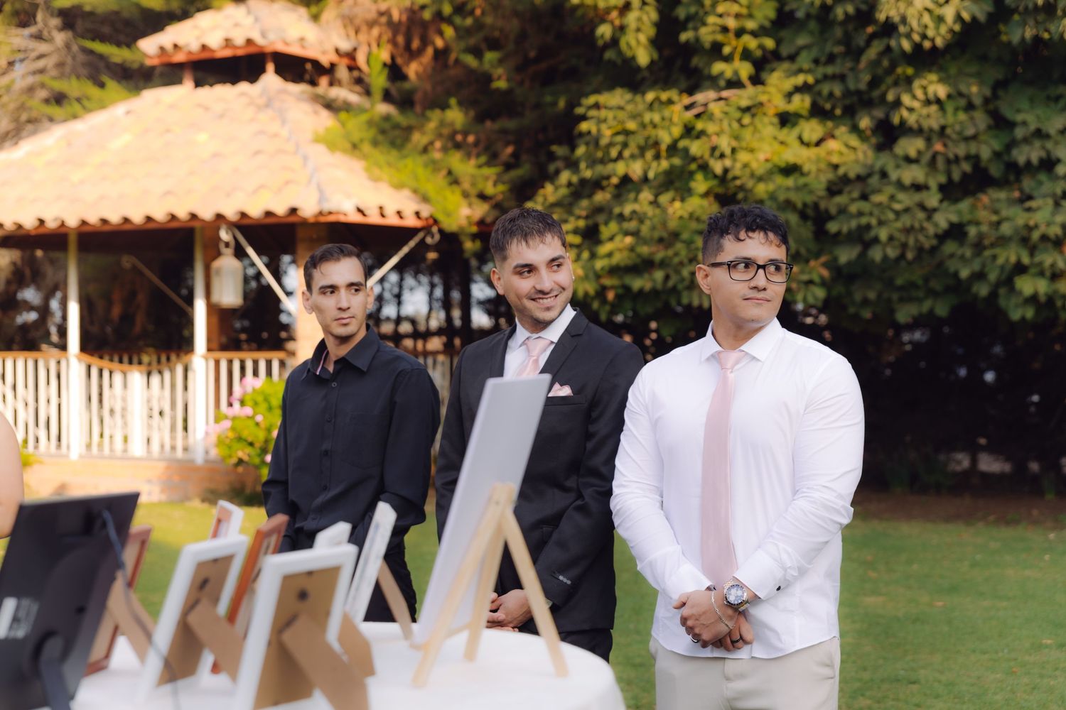 Three groomsmen stand outdoors at garden wedding ceremony with gazebo and lush greenery in background.