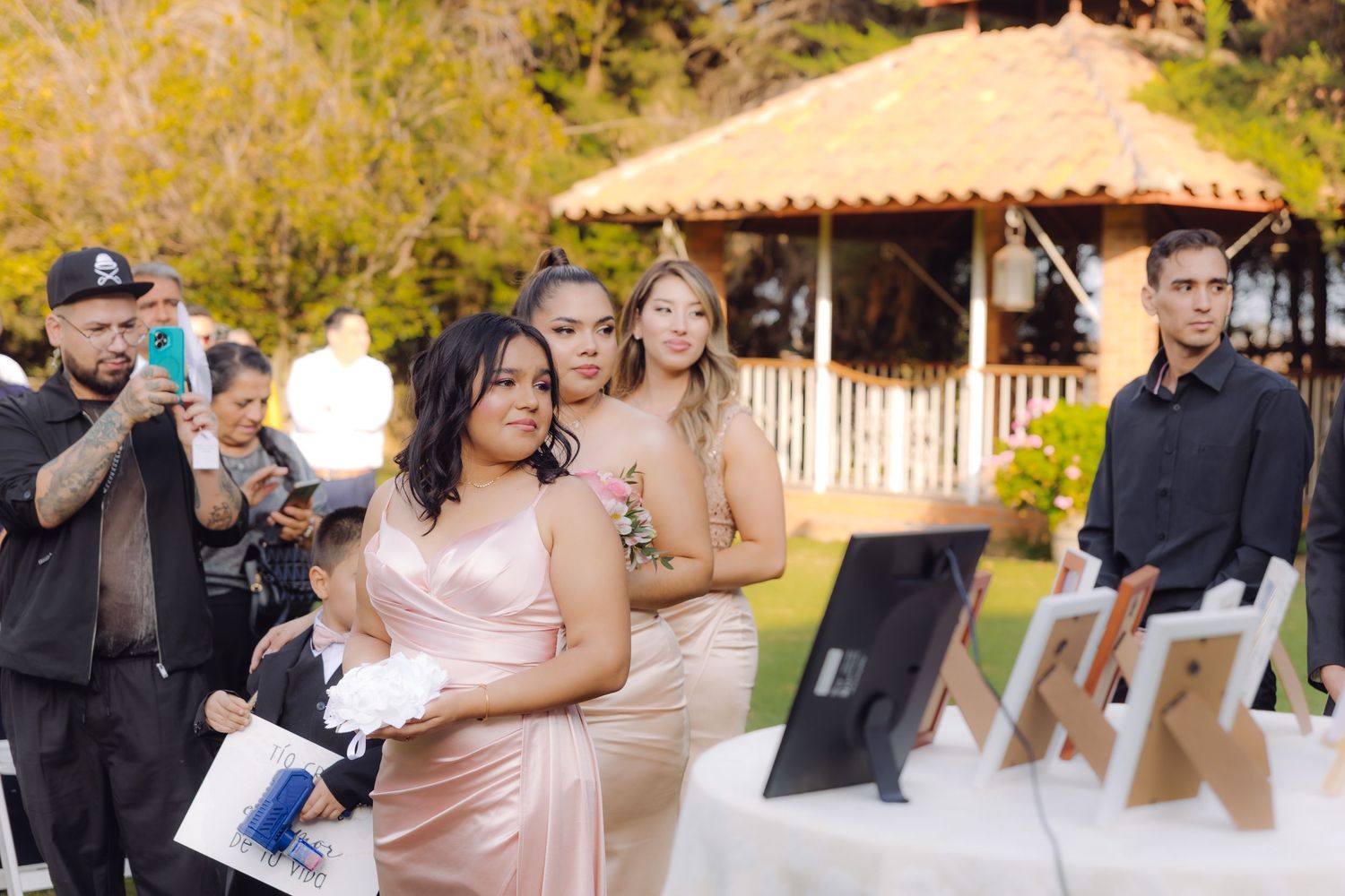 Bridesmaids in blush pink gowns walk down aisle at outdoor garden wedding ceremony with gazebo backdrop.