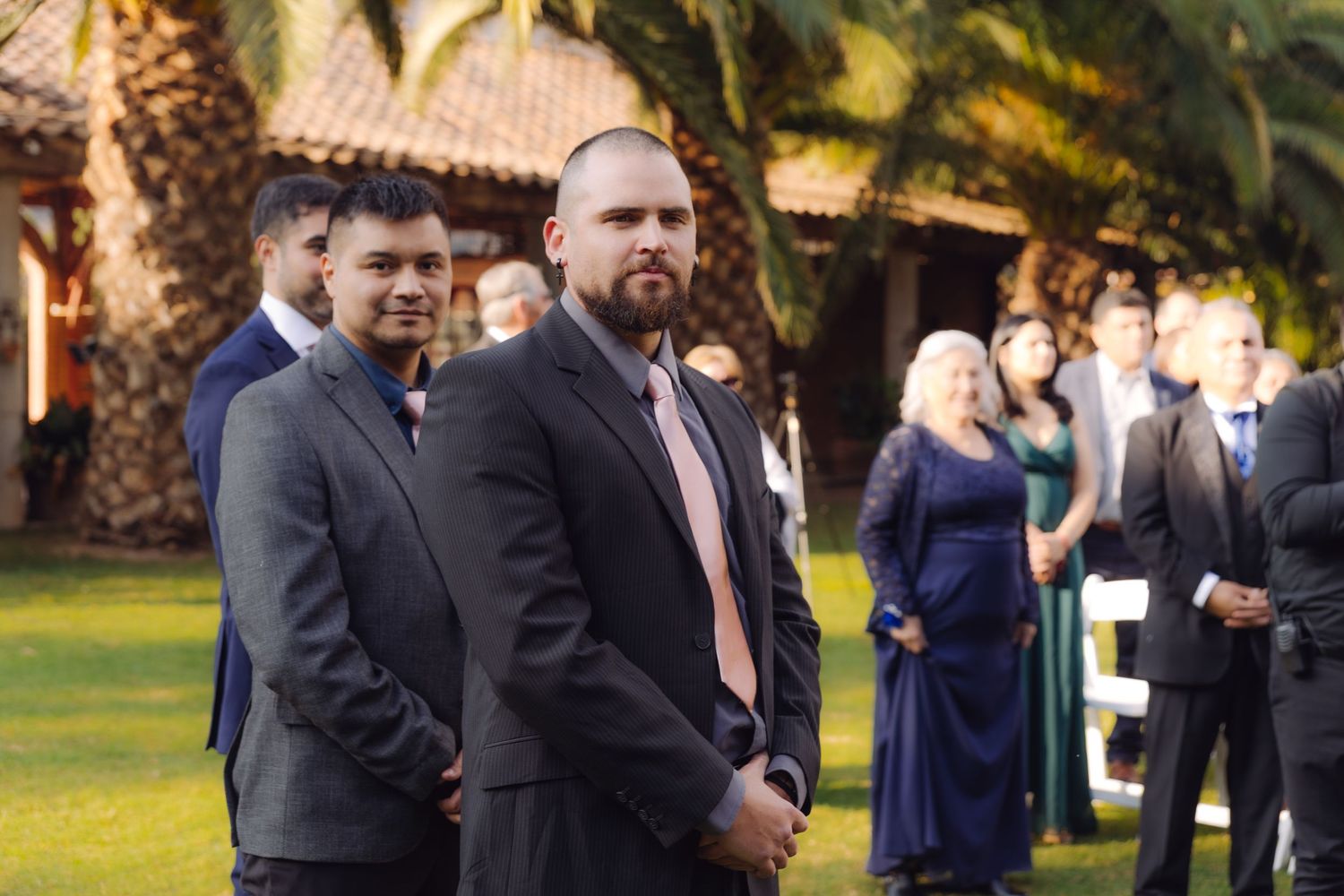 Two groomsmen in dark suits with pink ties stand attentively during outdoor wedding ceremony on green lawn.