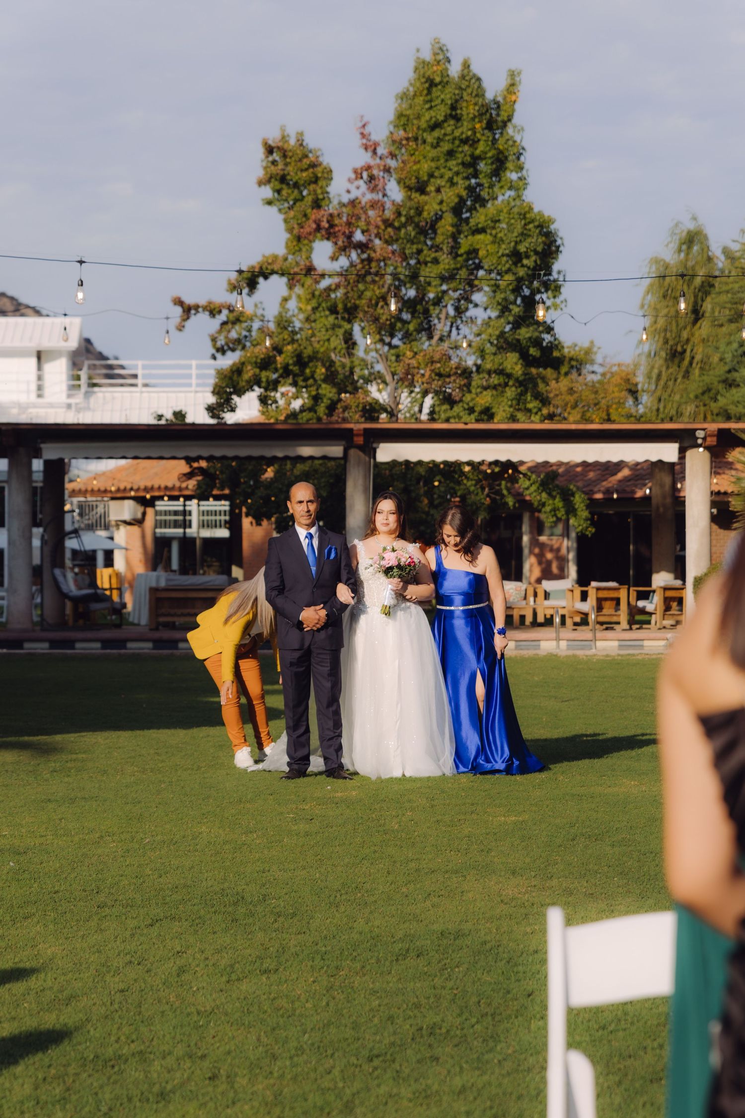 Bride in white gown walks down grassy aisle escorted by parents in blue outfits at outdoor wedding.