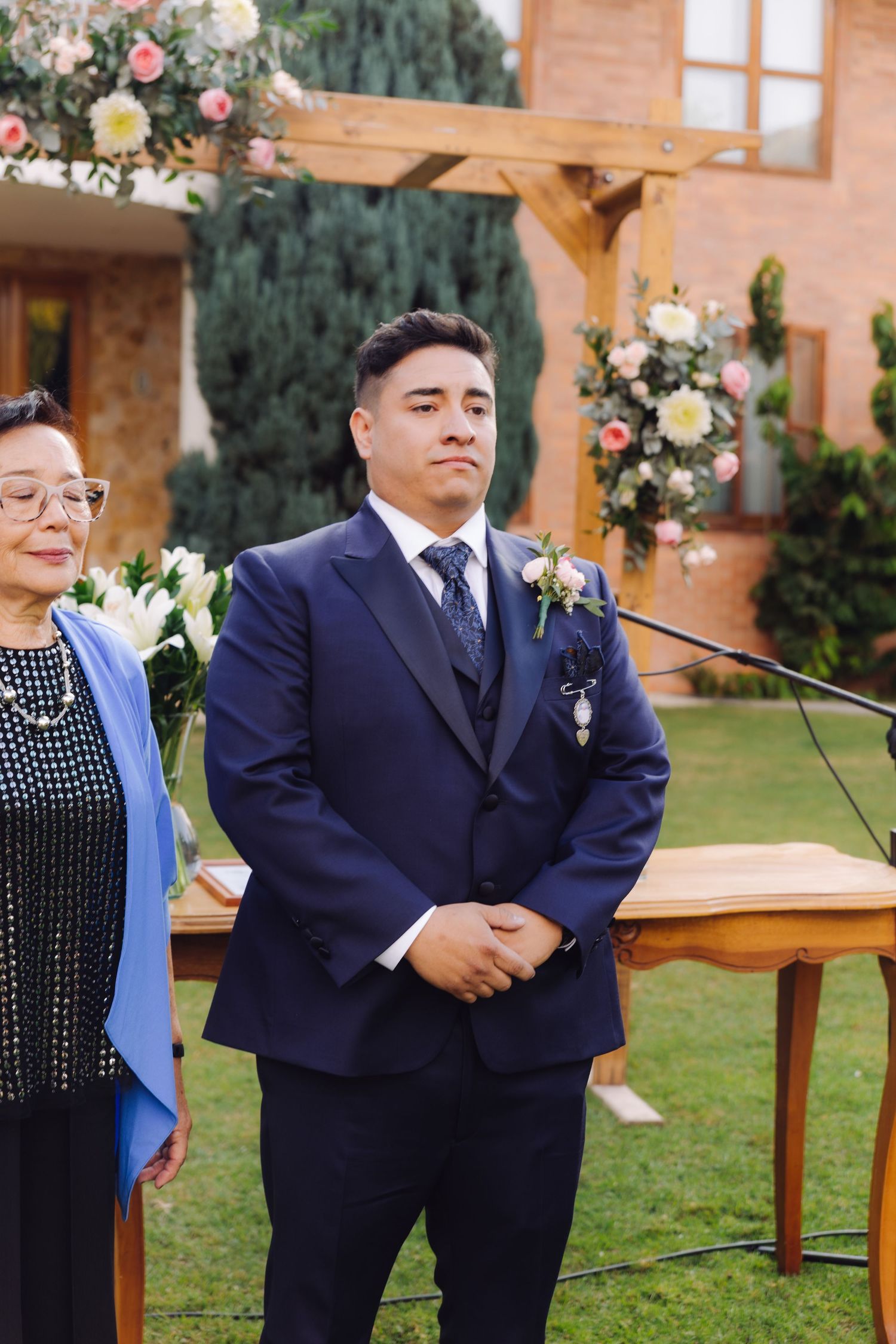 Groom in navy suit with floral boutonniere waits at outdoor wedding altar beside elderly woman in blue.
