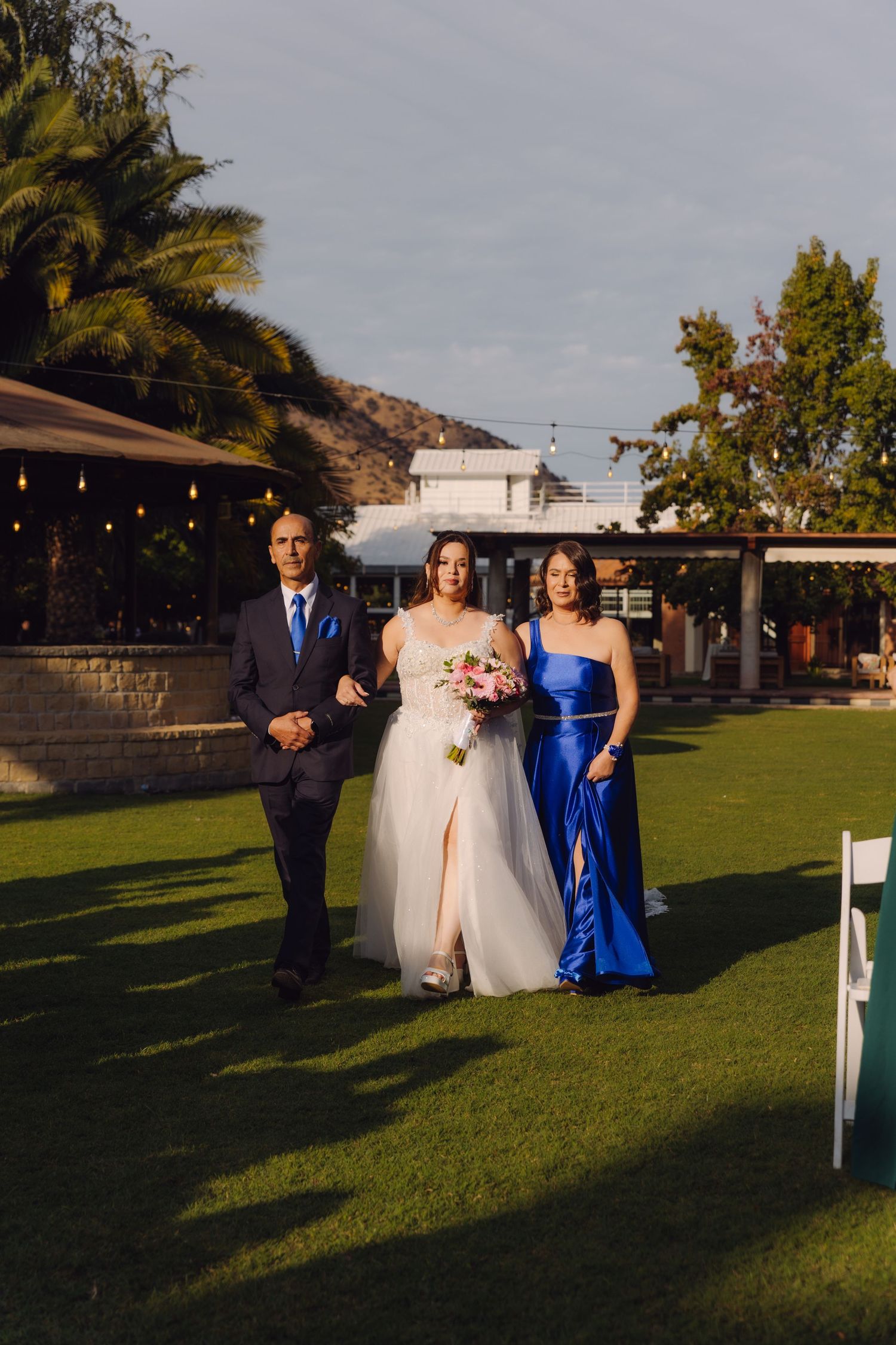 Bride in white gown with slit walks down grassy aisle holding bouquet, escorted by parents at sunset.
