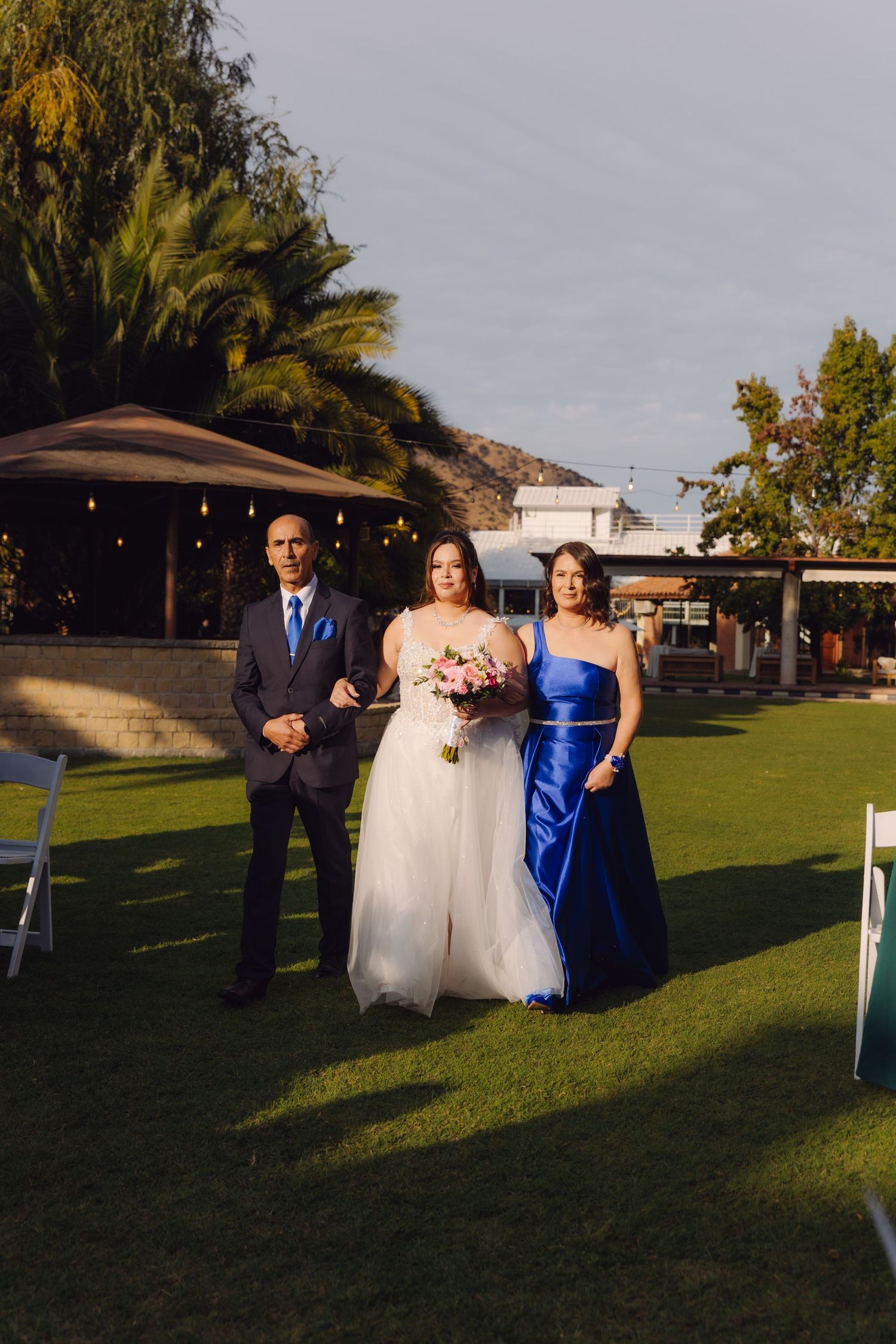 Bride holding pink bouquet walks down outdoor aisle flanked by parents in blue formal attire at sunset.