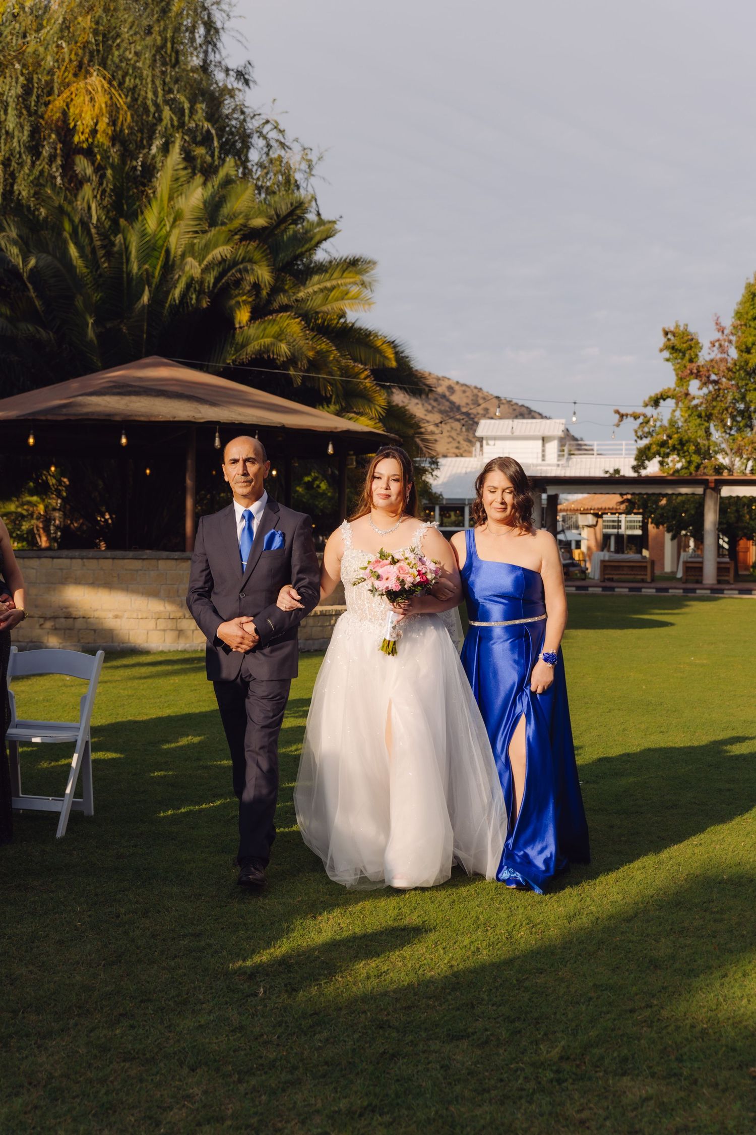 Bride in white gown walks down garden aisle with parents at golden hour outdoor wedding ceremony.