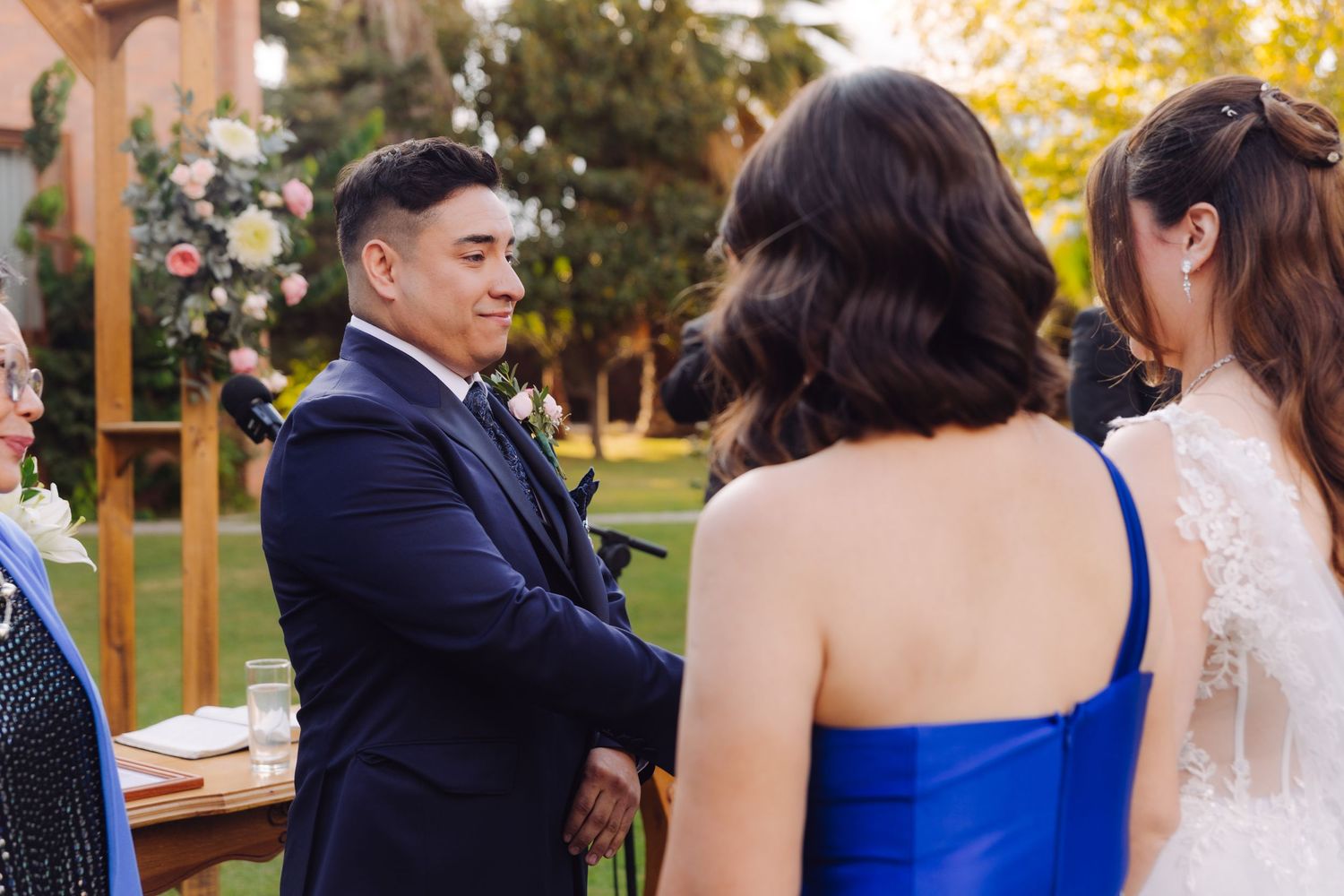 Groom in navy suit smiling at bride during outdoor wedding ceremony on green lawn.