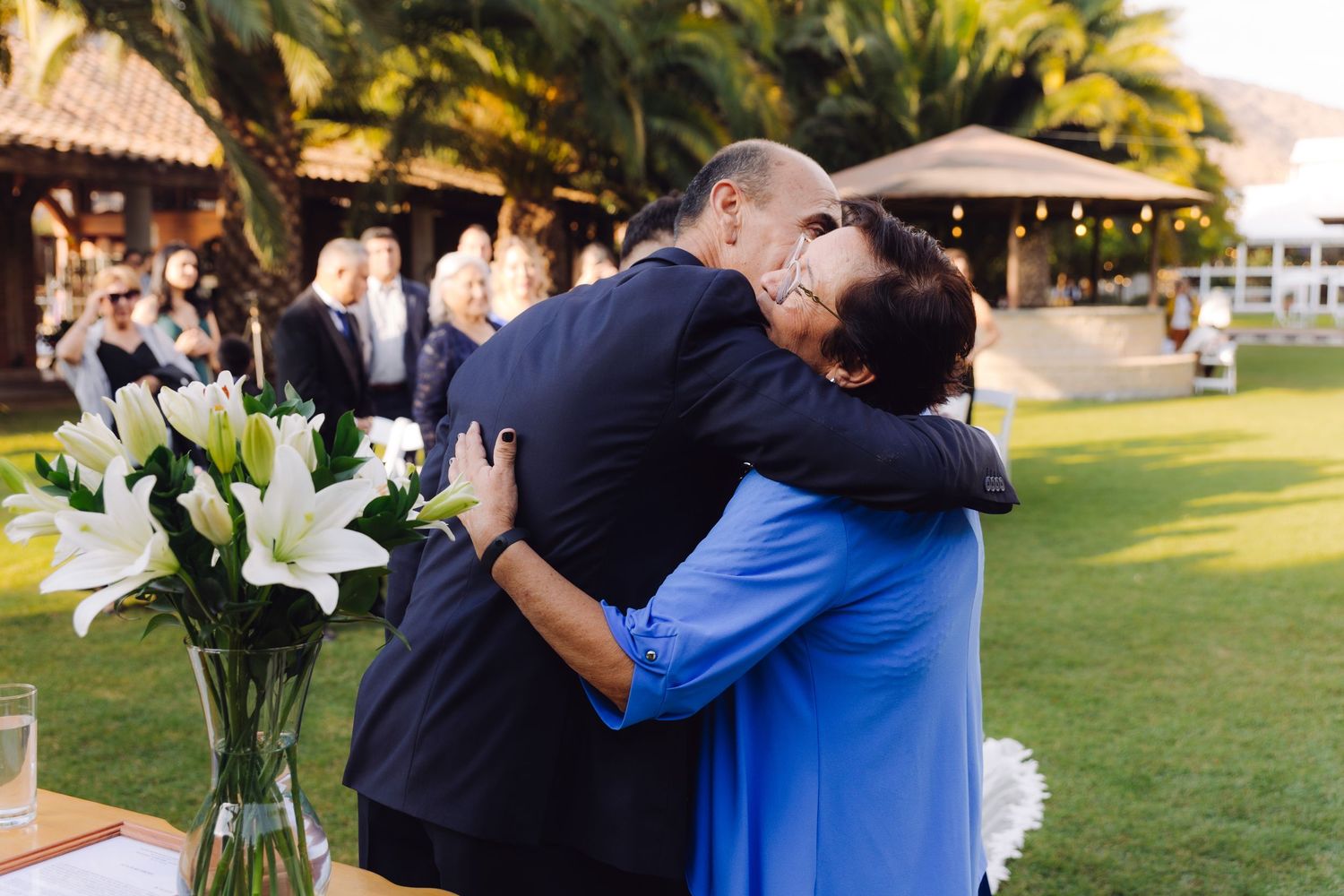 Wedding guests sharing warm embrace near floral arrangement with white lilies outdoors.