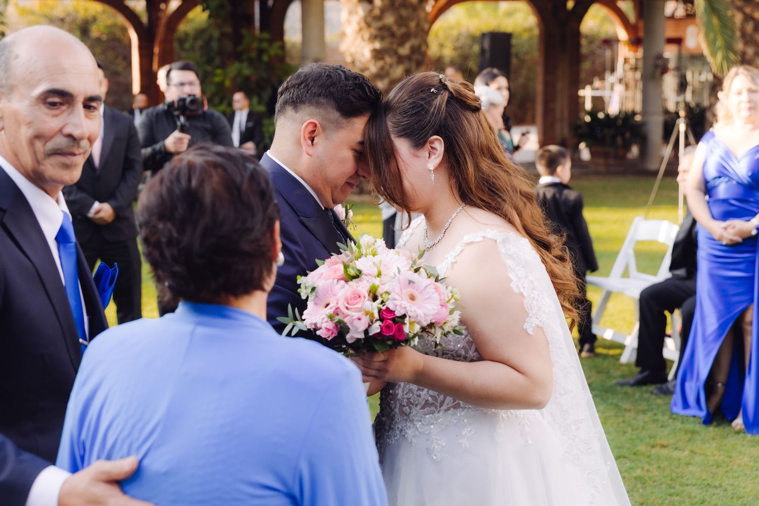Bride in lace gown holding pink bouquet sharing tender moment with groom and guest.