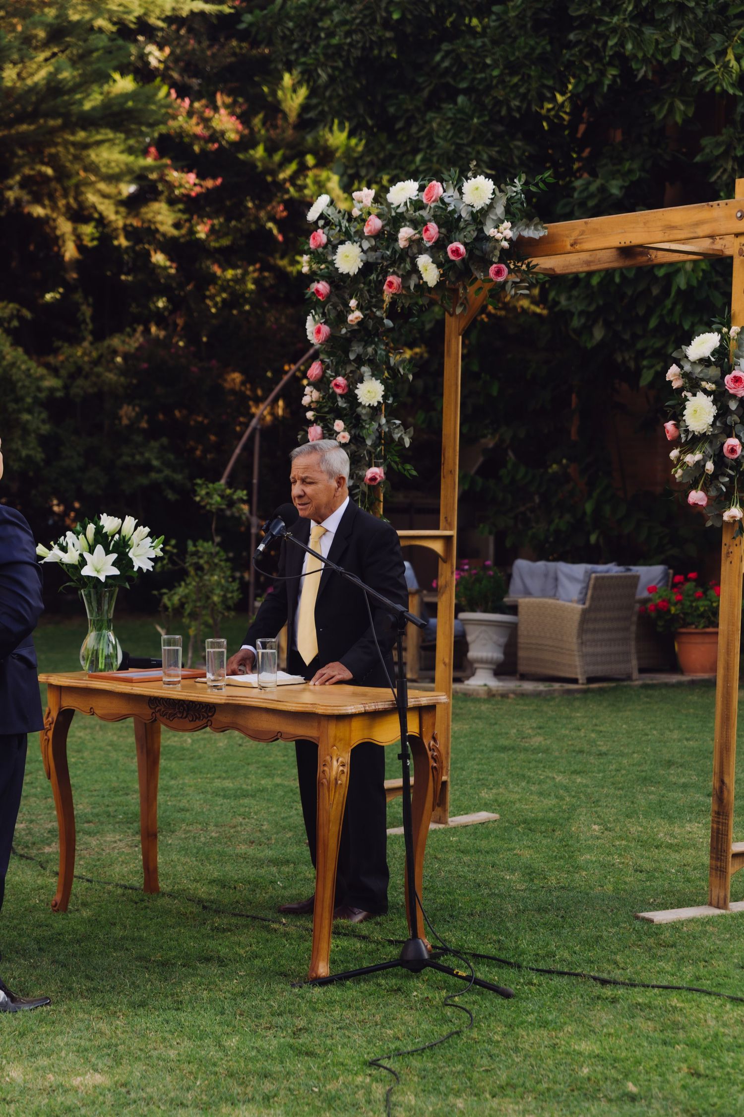 Wedding officiant speaks at a wooden altar decorated with pink and white flowers during an outdoor ceremony.