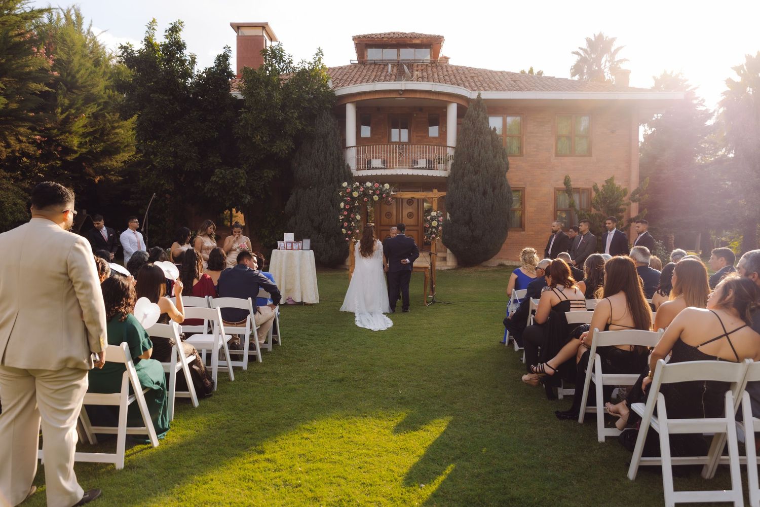 Outdoor wedding ceremony on a green lawn with white chairs and a grand brick mansion backdrop.