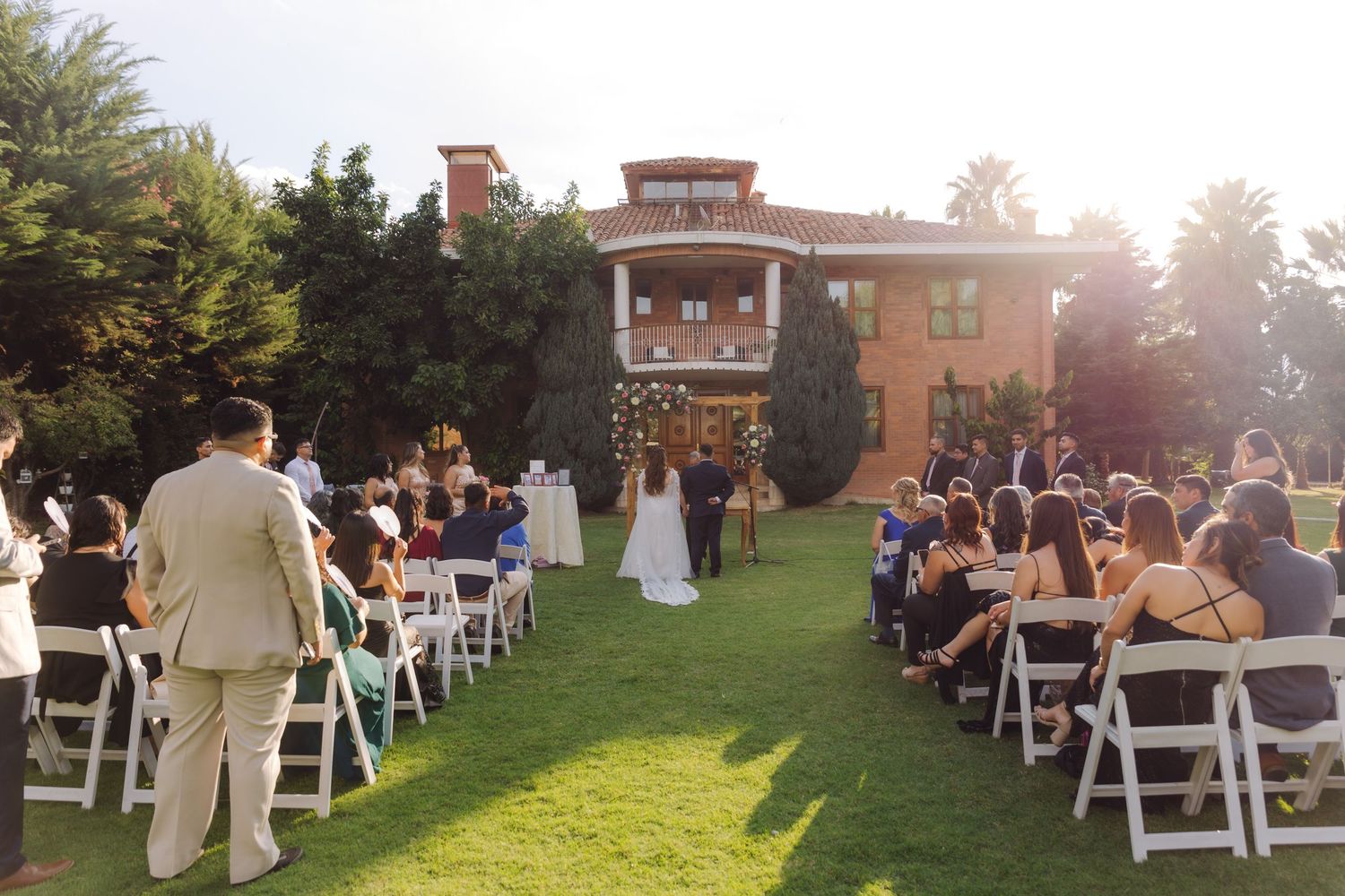 Guests seated in white chairs witness a wedding ceremony on a sunlit lawn in front of an elegant estate.