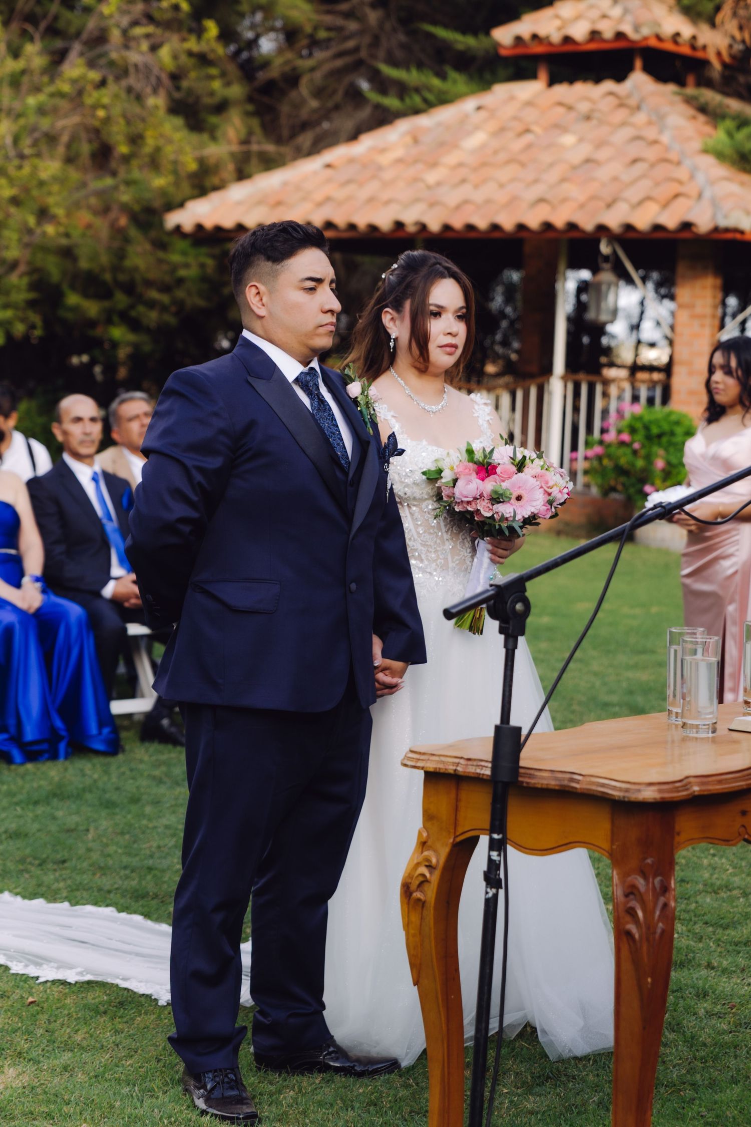 Bride holding a pink bouquet and groom in navy suit stand at the altar during an outdoor wedding ceremony.
