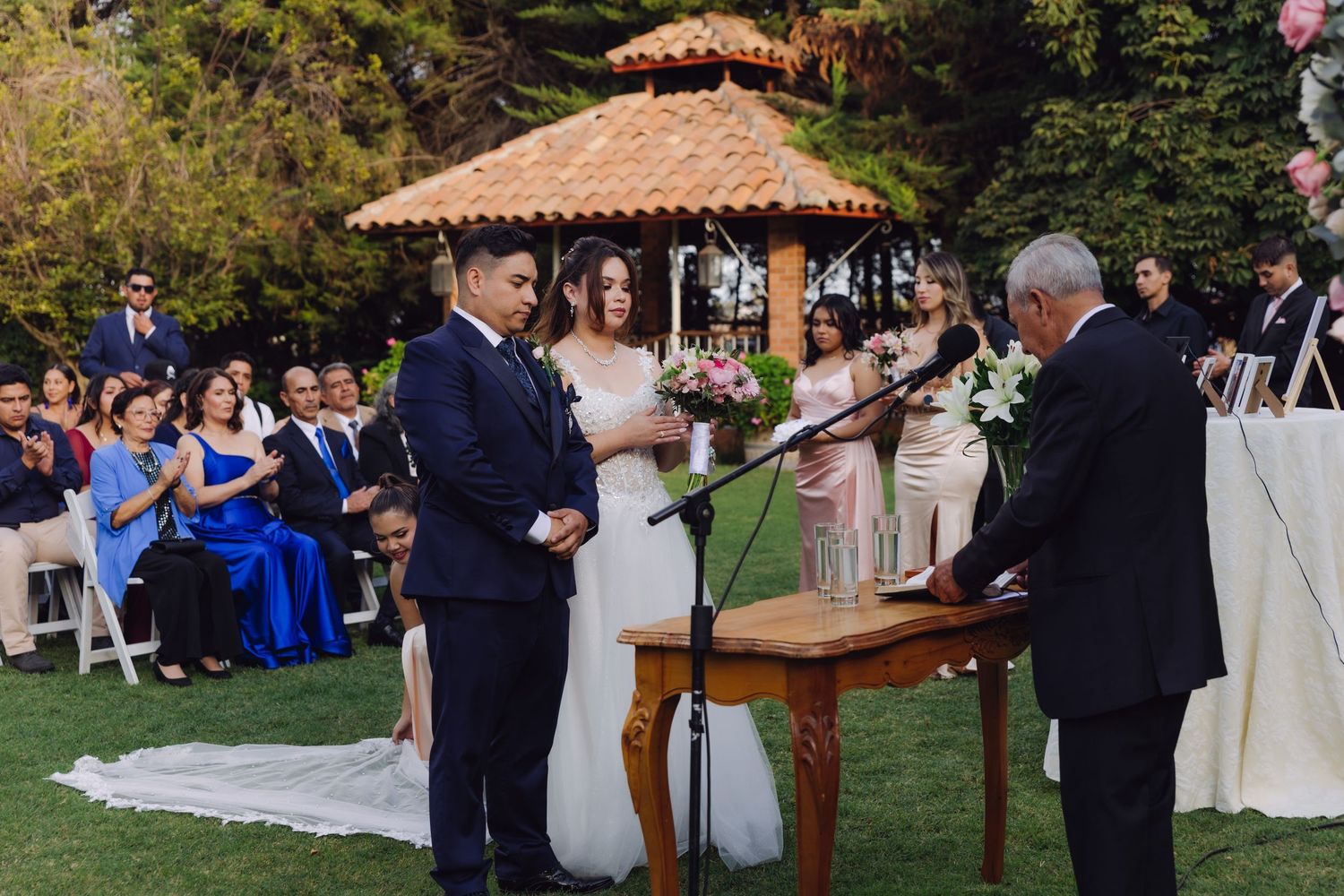 Bride and groom face each other smiling during their outdoor garden wedding ceremony with guests applauding.